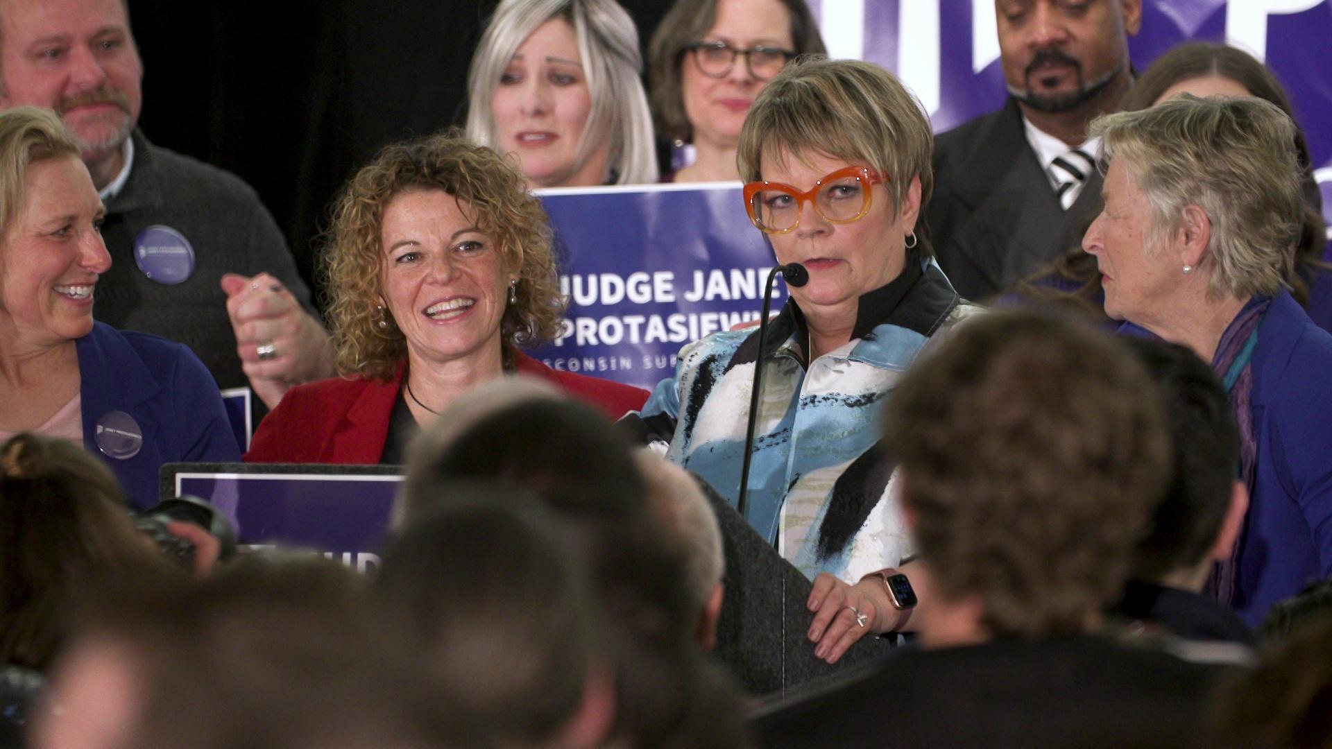 A still image from a video shows Jill Karofsky, Rebecca Dallet, Janet Protasiewicz and Ann Walsh Bradley standing among a crowd of people, with several holding 'Judge Janet Protasiewicz' campaign signs.
