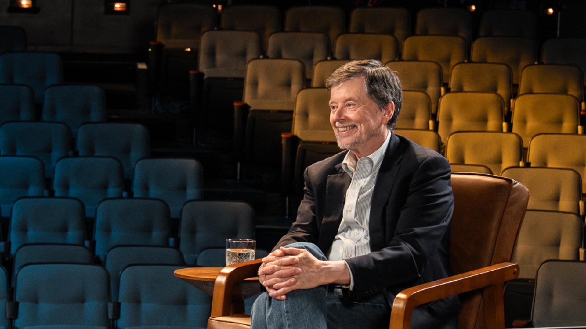 Filmmaker Ken Burns sitting in a leather chair with auditorium seating in the background.