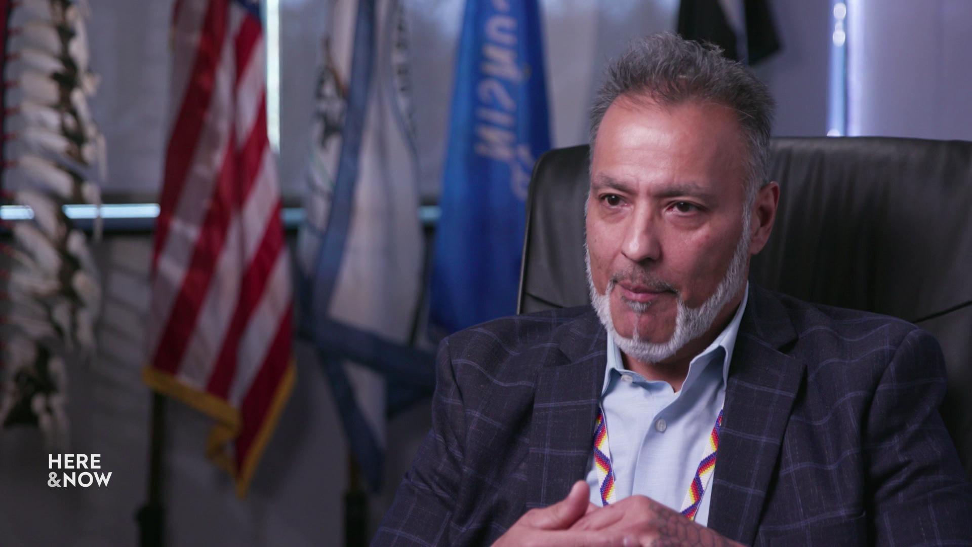 Jon Greendeer sits in a high-back chair with flags in the background.