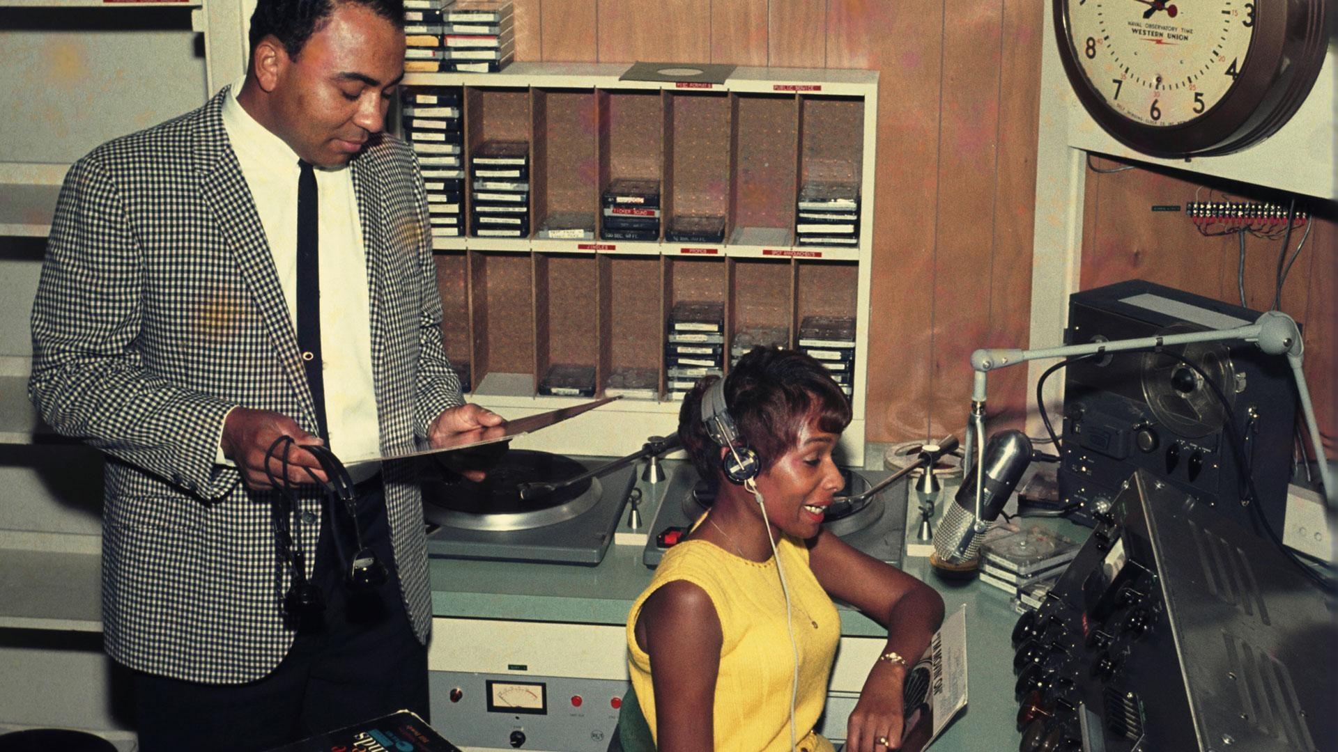 Archival photo of man and woman in studio with casette tapes and records.