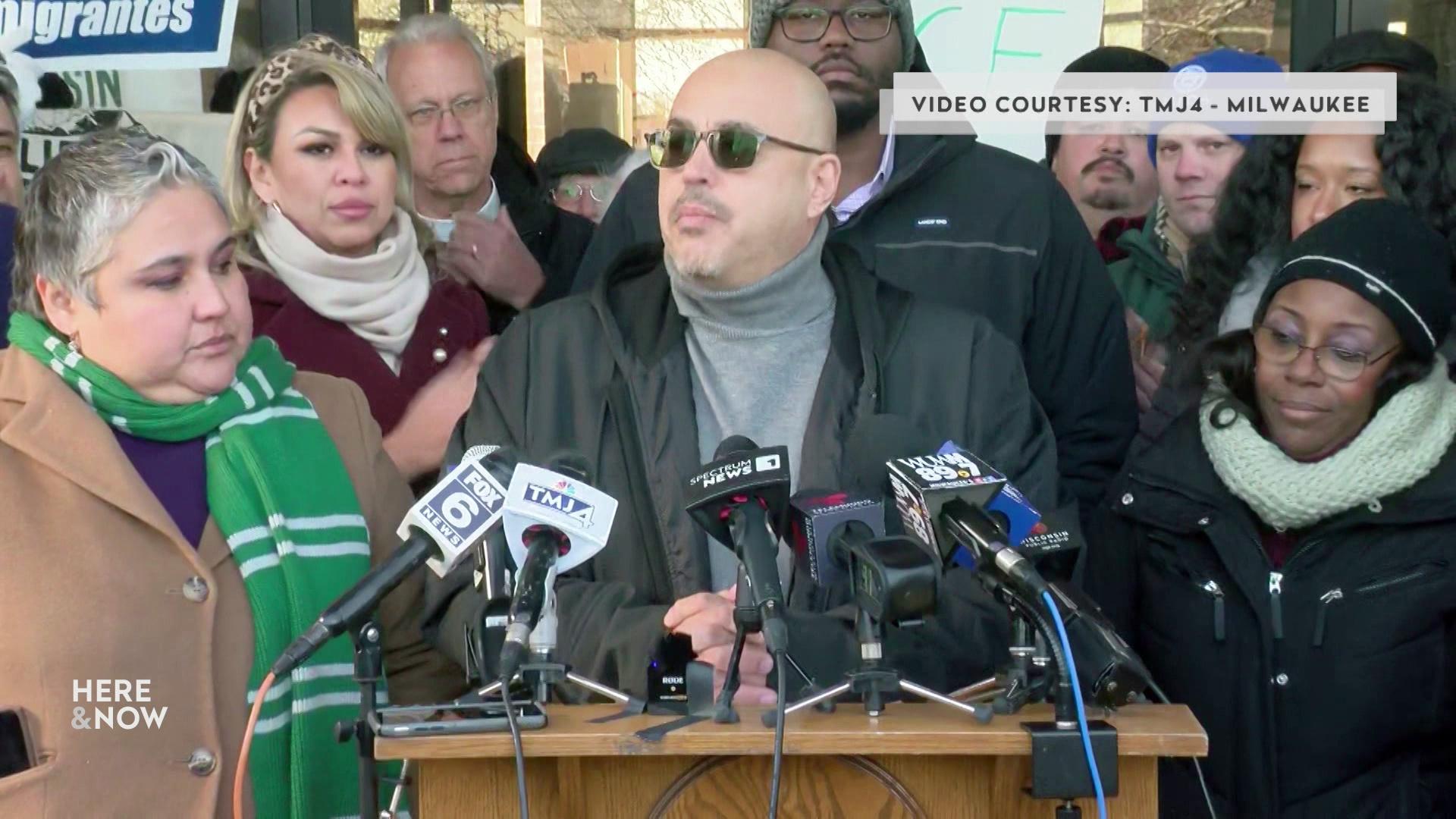 A still image from a video shows José Pérez speaking into multiple microphones with various media flags on a podium with people standing around him with a graphic at top reading 'Video Courtesy: TMJ4 - Milwaukee.'