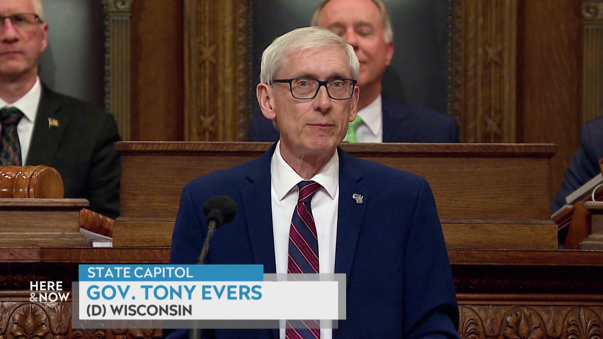 A still image from a video shows Tony Evers speaking into a microphone in front of three individuals seated on a dais, with a graphic at bottom reading 'Gov. Tony Evers' and '(D) Wisconsin.'