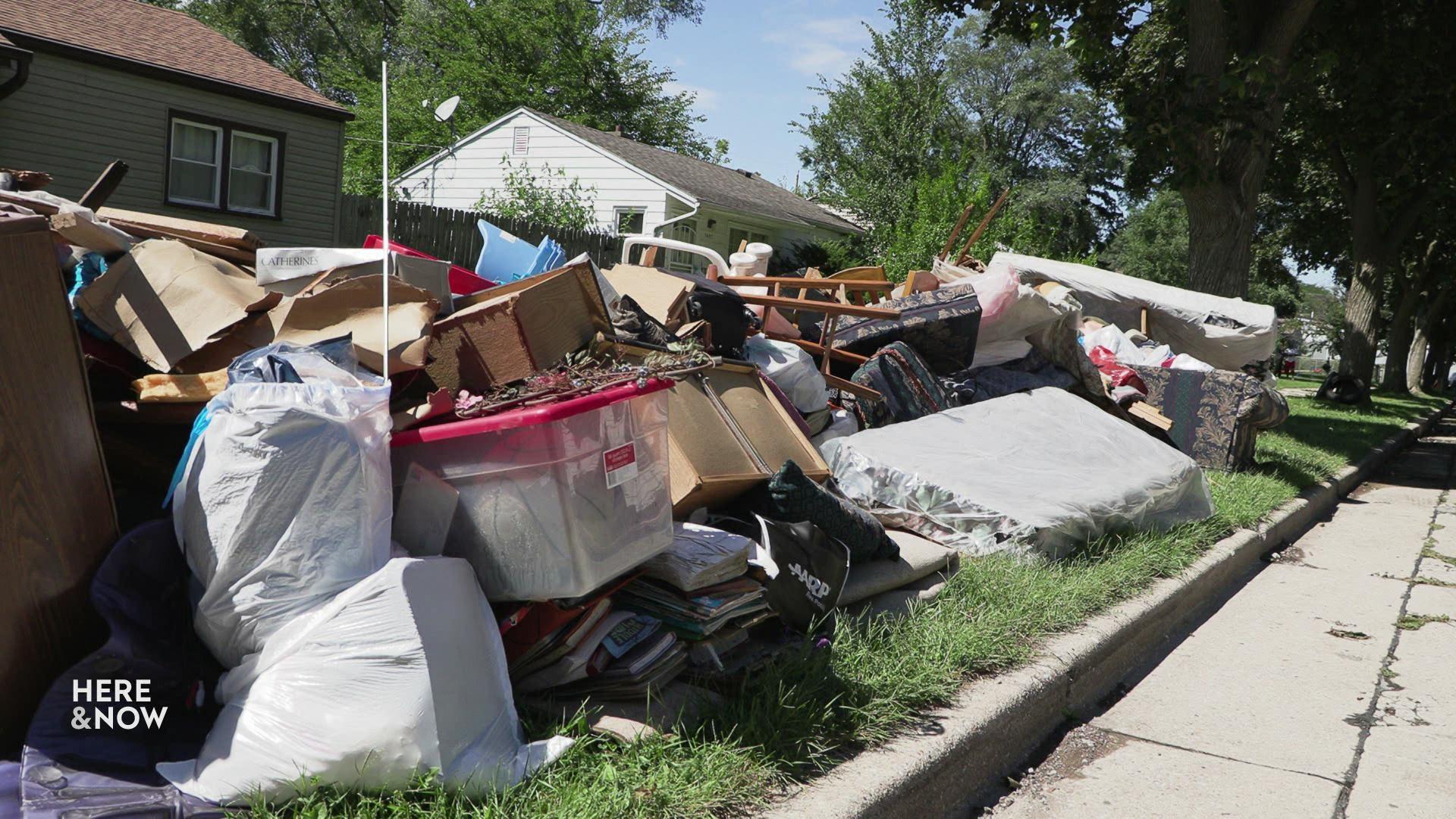 A still image shows piles of flood-damaged housewares stacked up on the side of the road, including mattresses, bins and furniture.