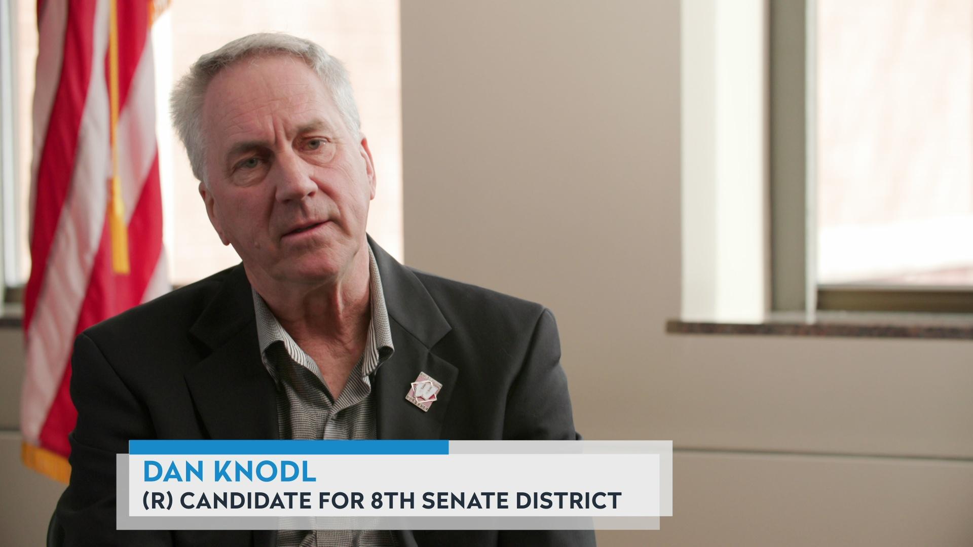 A still image from a video shows Dan Knodl sitting in a room with windows and a U.S. flag in the background, with a graphic at bottom reading 'Dan Knodl' and '(R) Candidate for 8th Senate District.' 