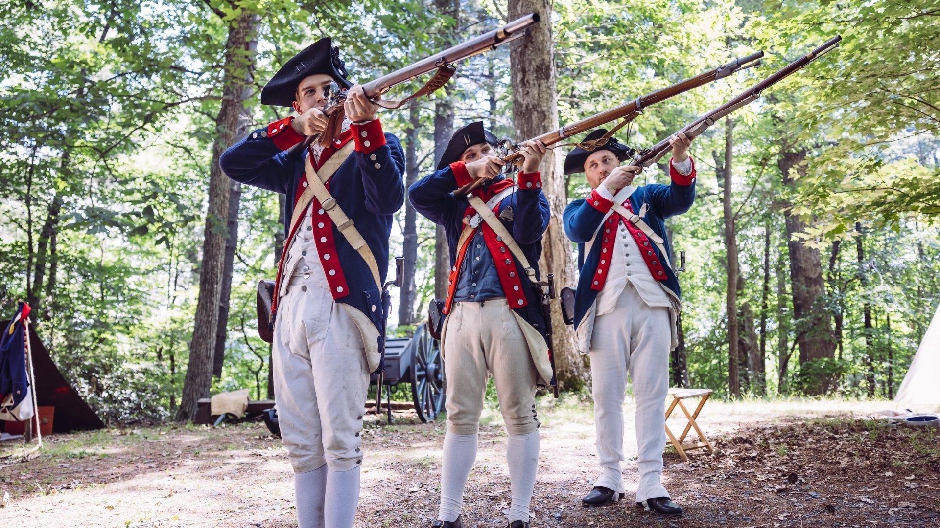 Three white men dressed in revolutionary solder uniforms and holding up rifles.