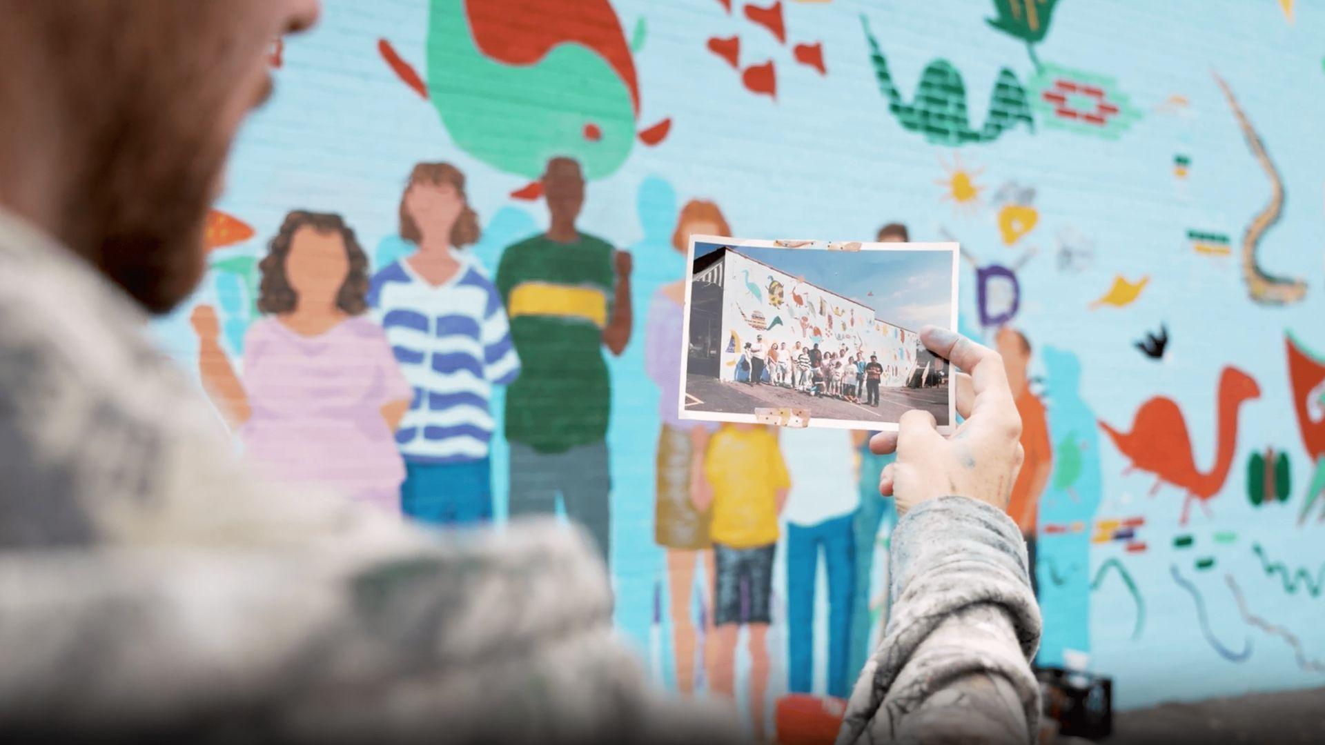 A person holding up an image of a mural in front of that mural.