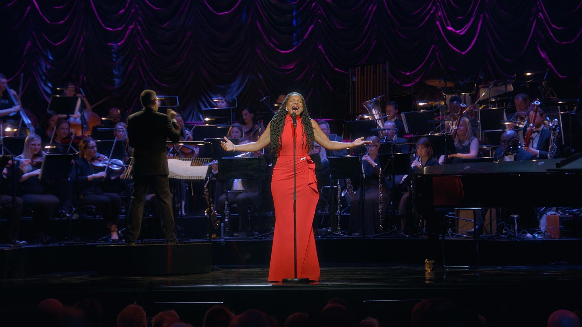 Audra McDonald, wearing a red dress, and singing on stage in front of an orchestra