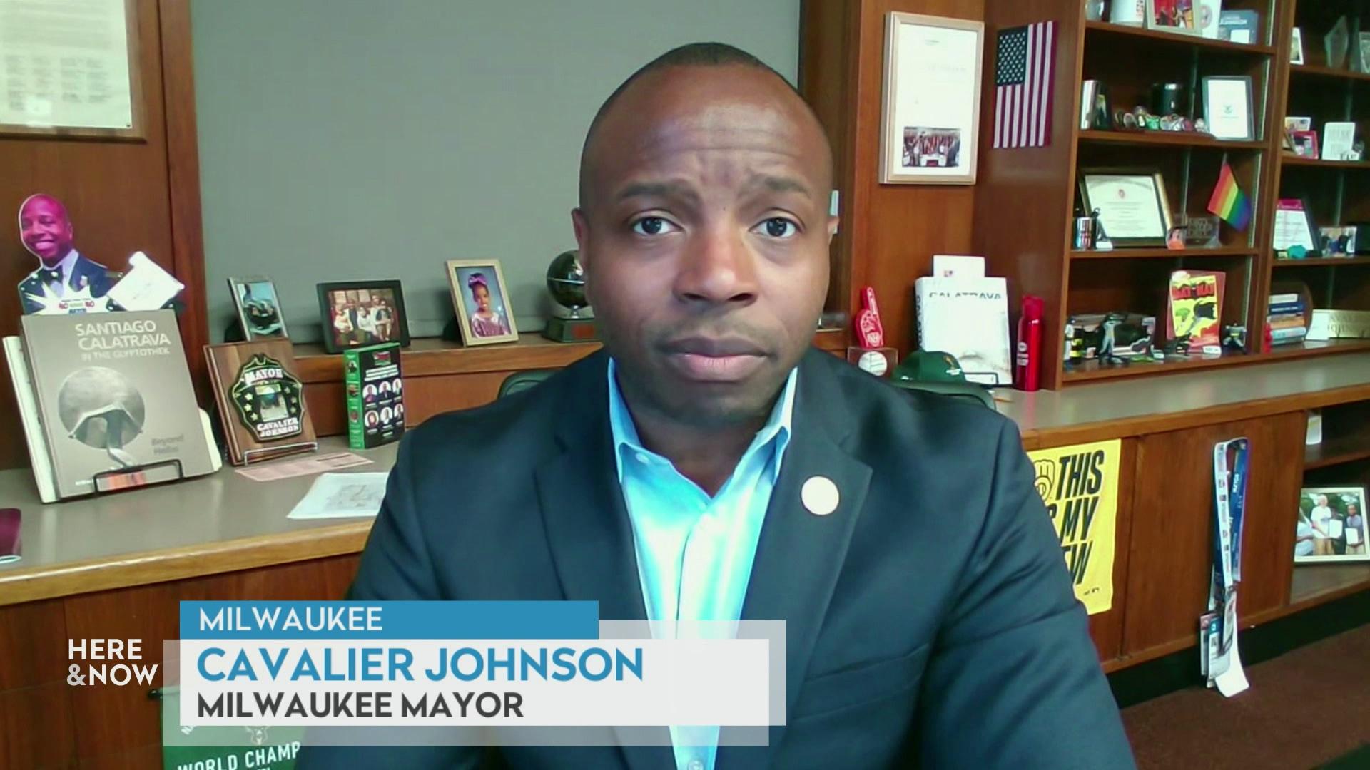 A still image from a video shows Mayor Cavalier Johnson seated in front of wood paneled cabinets and shelves with a graphic at bottom reading 'Milwaukee,' 'Cavalier Johnson' and 'Milwaukee Mayor.'
