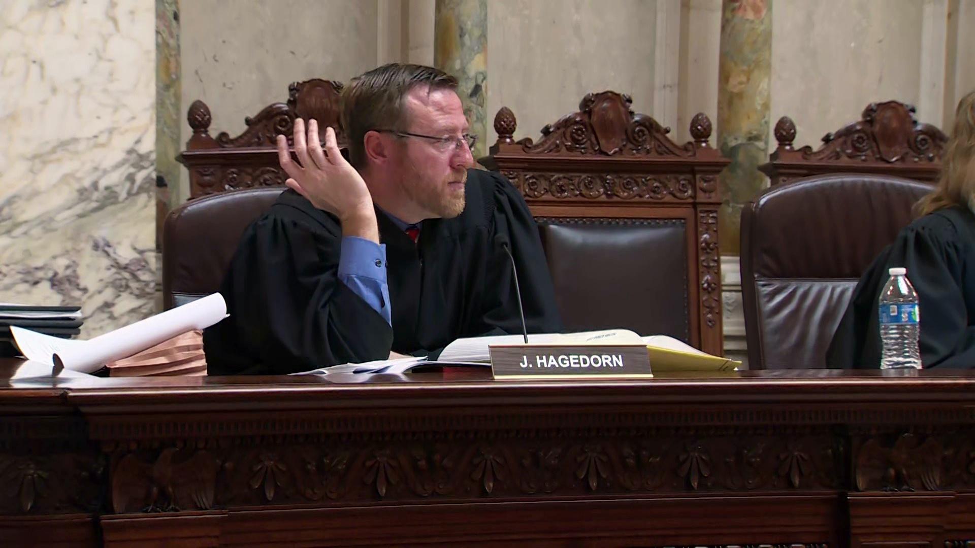 A still image from a video shows Brian Hagedorn holding his right hand up while seated in a high-backed chair behind a wooden dais in a room with marble walls and pillars.