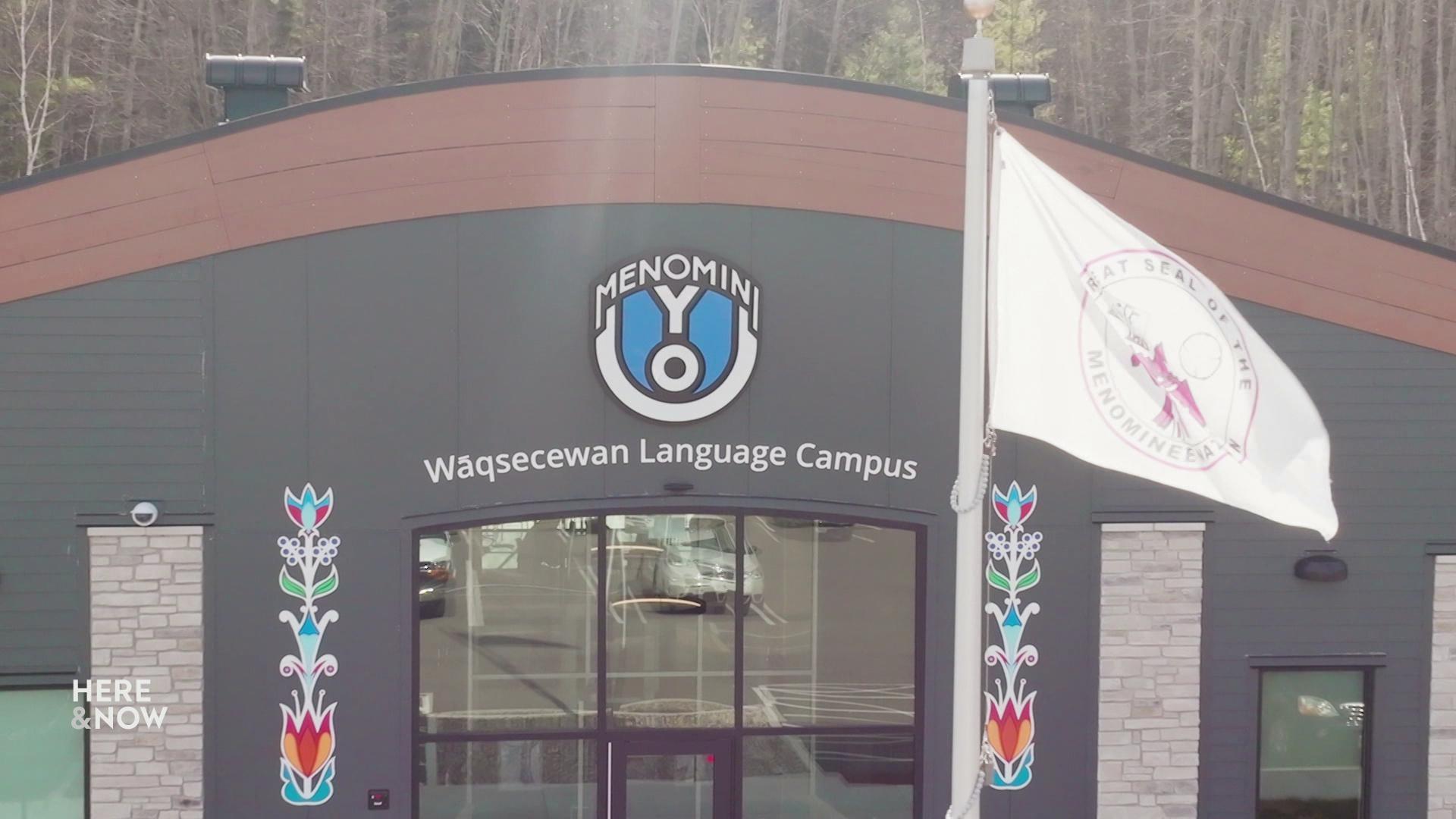 A still image shows the Menomini yoU language campus, a wide arched gray building with floral designs on the sides of a large glass entrance with a Menominee Nation flag waving in front of it.