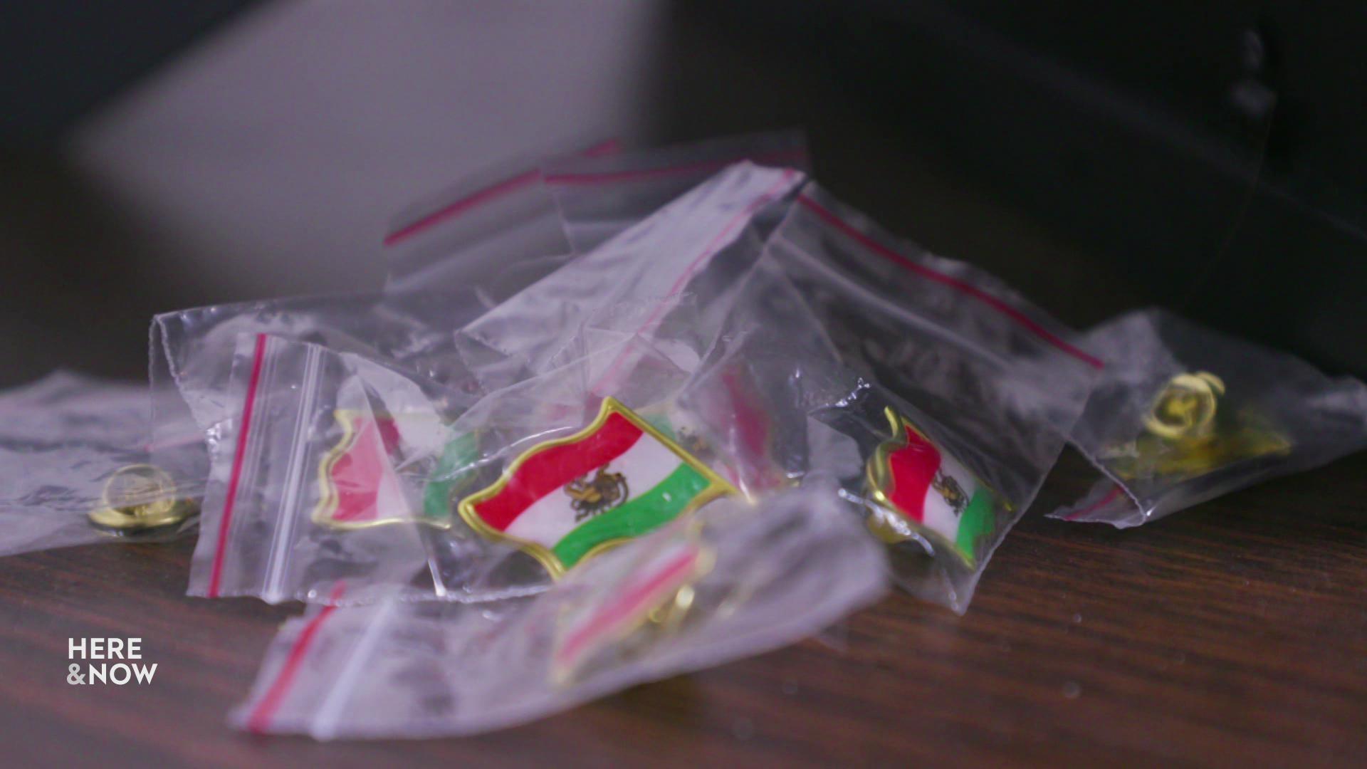 A still image shows a pile of Iranian flag pins inside small plastic bags on a wooden table.