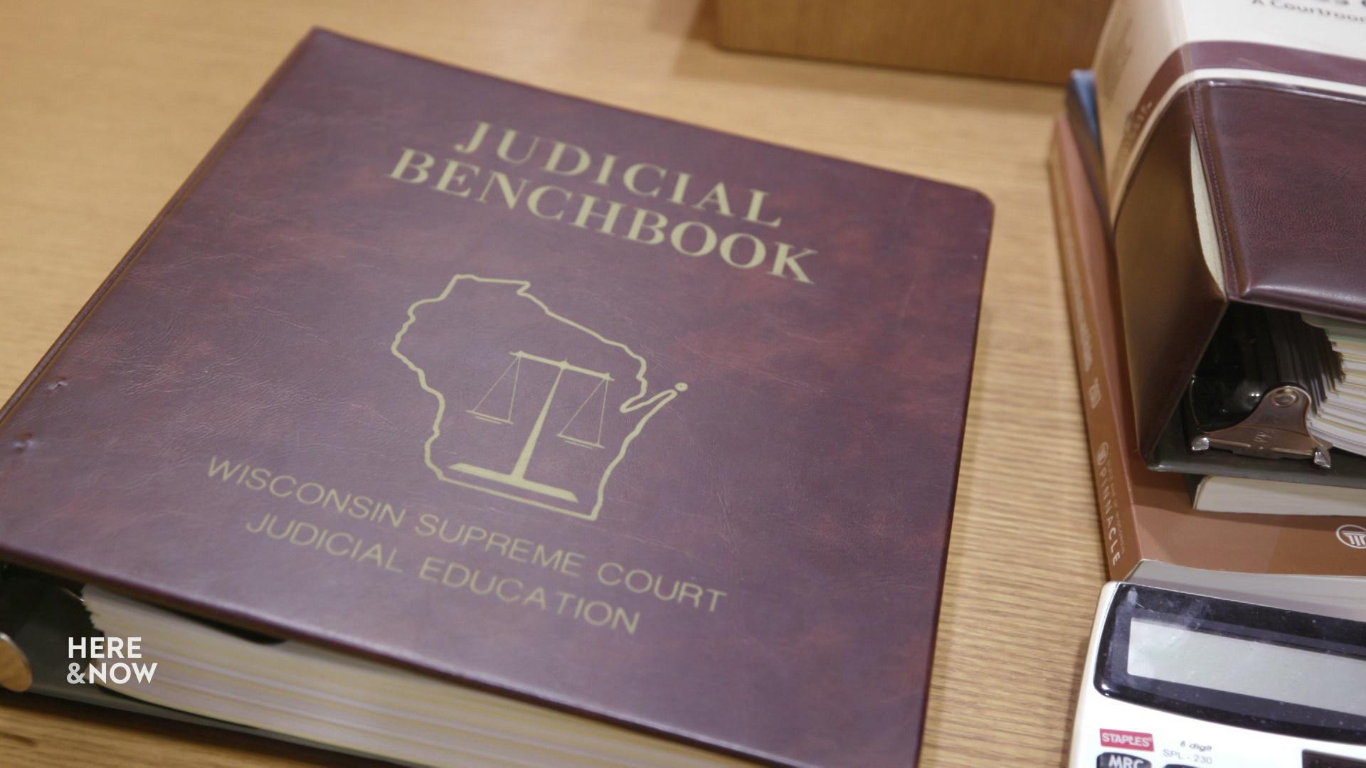 A still image from a video shows a book with a cover featuring an outline of Wisconsin and the words 'Judicial Benchbook' and 'Wisconsin Supreme Court' and 'Judicial Education.'