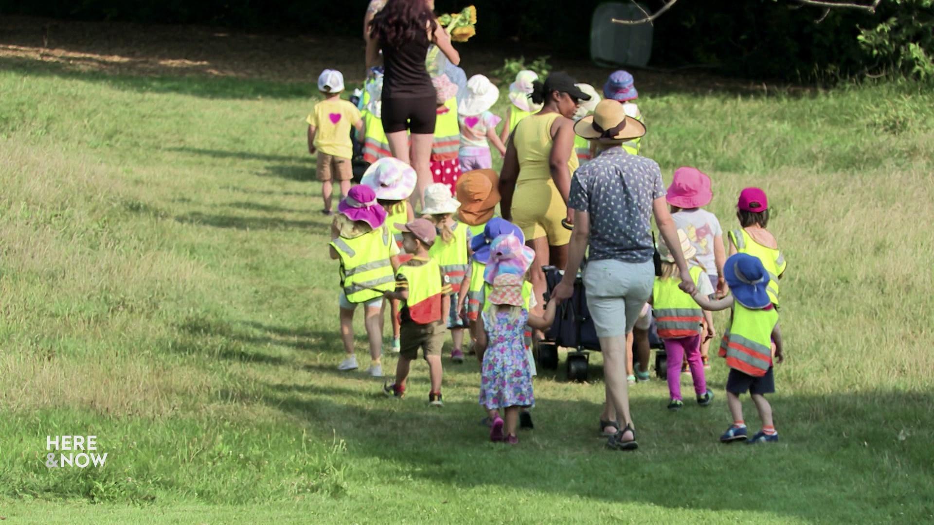 A still image shows a group of children wearing neon safety vests and sun hats while holding hands with adults as they walk through a green field.