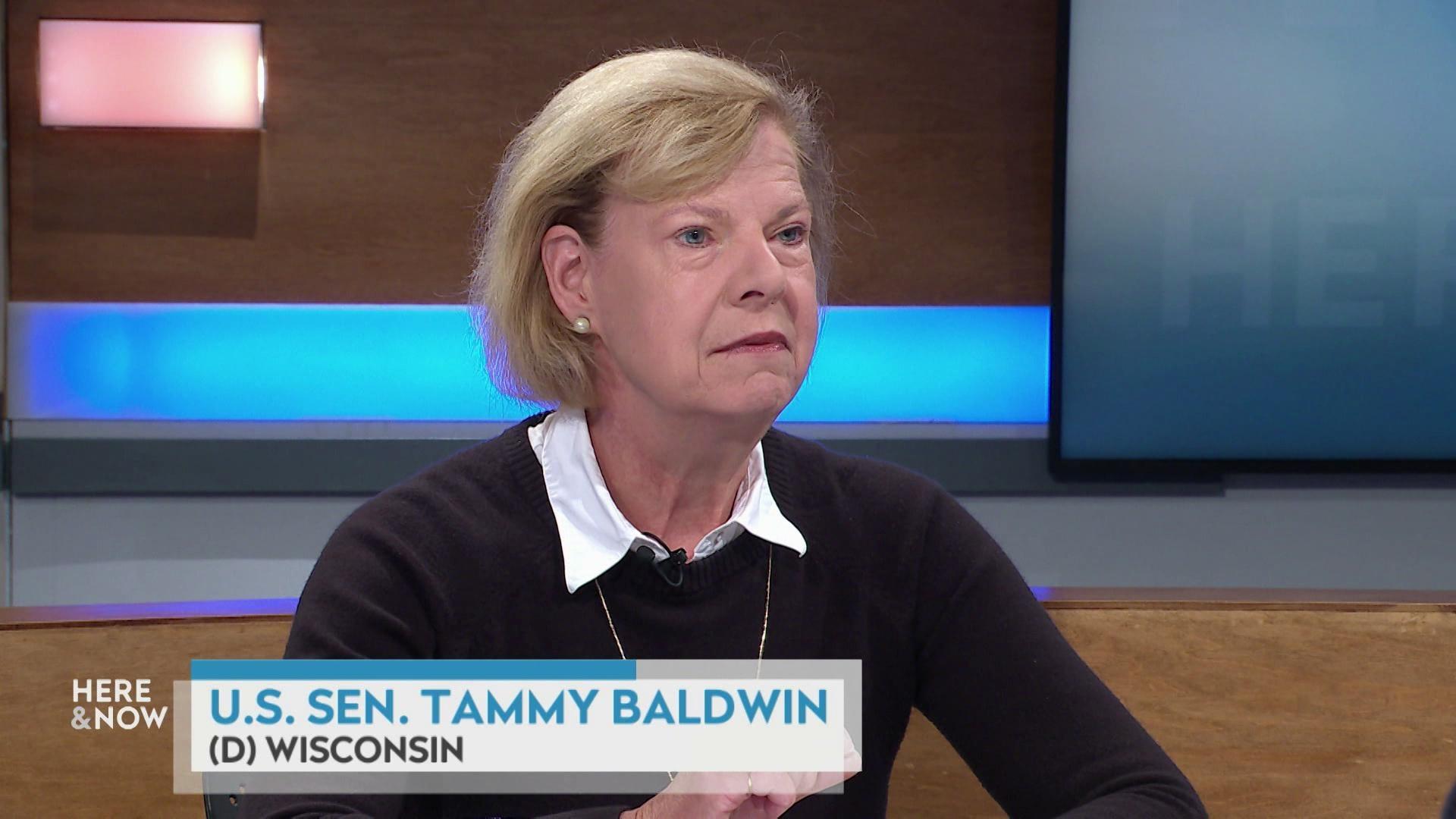 A still image shows Tammy Baldwin seated at the 'Here & Now' set featuring wood paneling, with a graphic at bottom reading 'U.S. Sen. Tammy Baldwin' and '(D) Wisconsin'