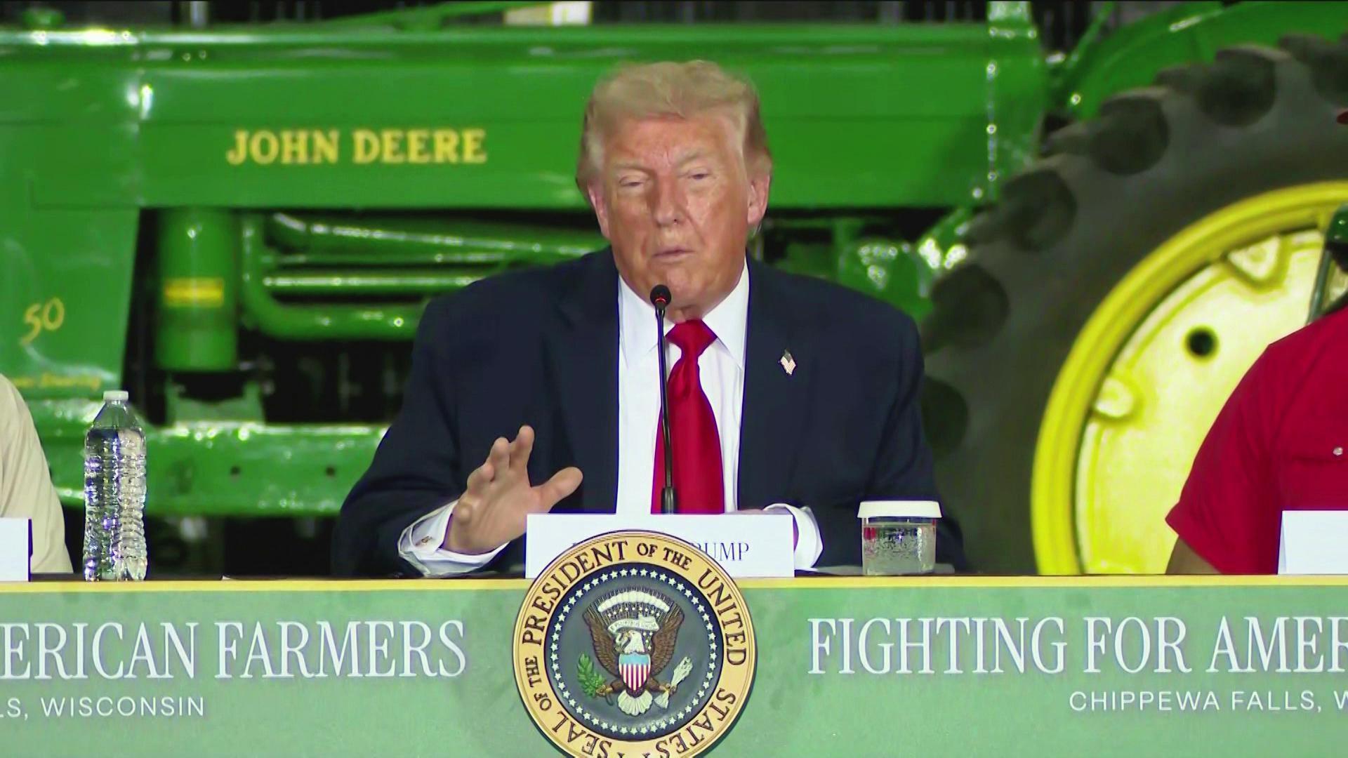 A still image shows President Donald Trump seated at bannered table with the Seal of the President of the United States on the front and graphics on either side that read FIGHTING FOR AMERICAN FARMERS and Chippewa Falls, Wisconsin.