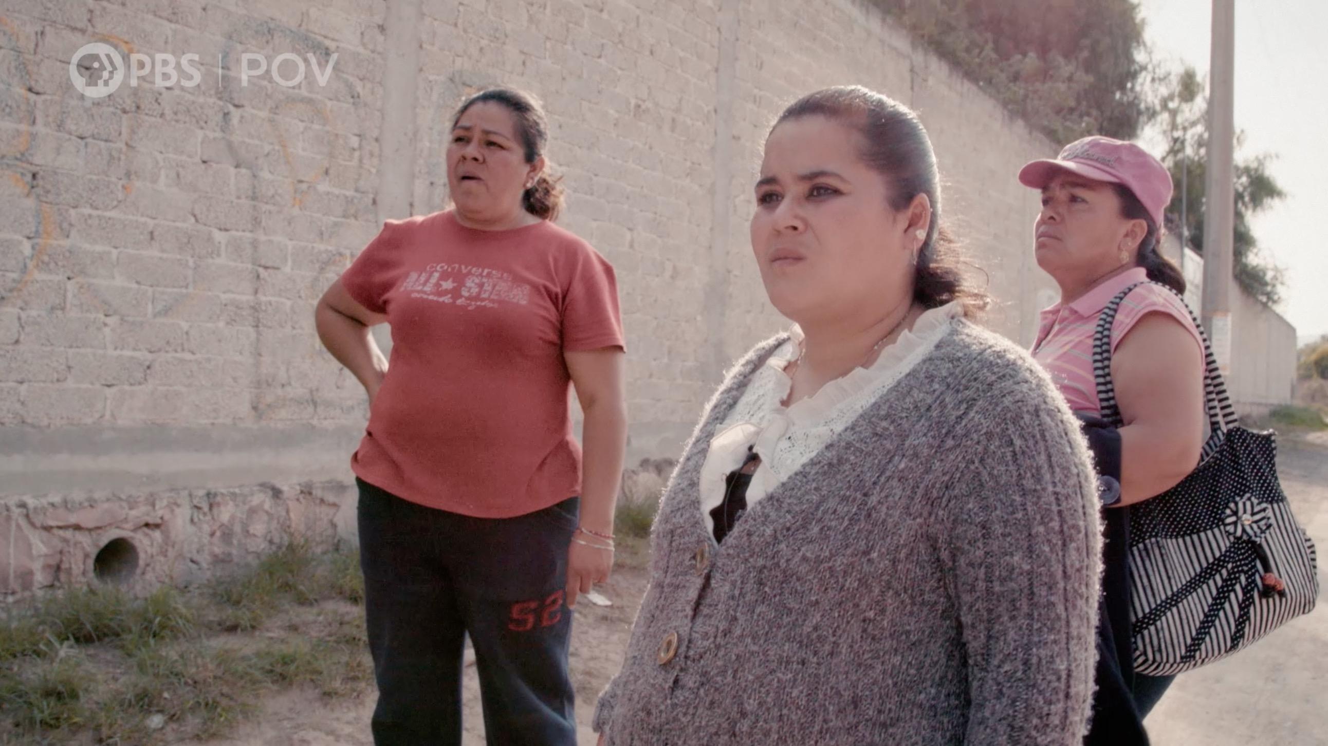 Three women stand outside with a white brick wall behind them.