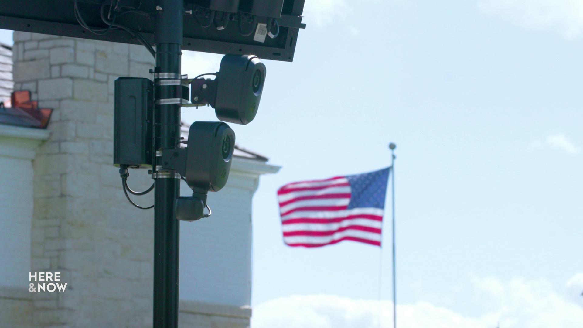 A still image shows a flock camera secured to a black pole with an American flag waving behind it.