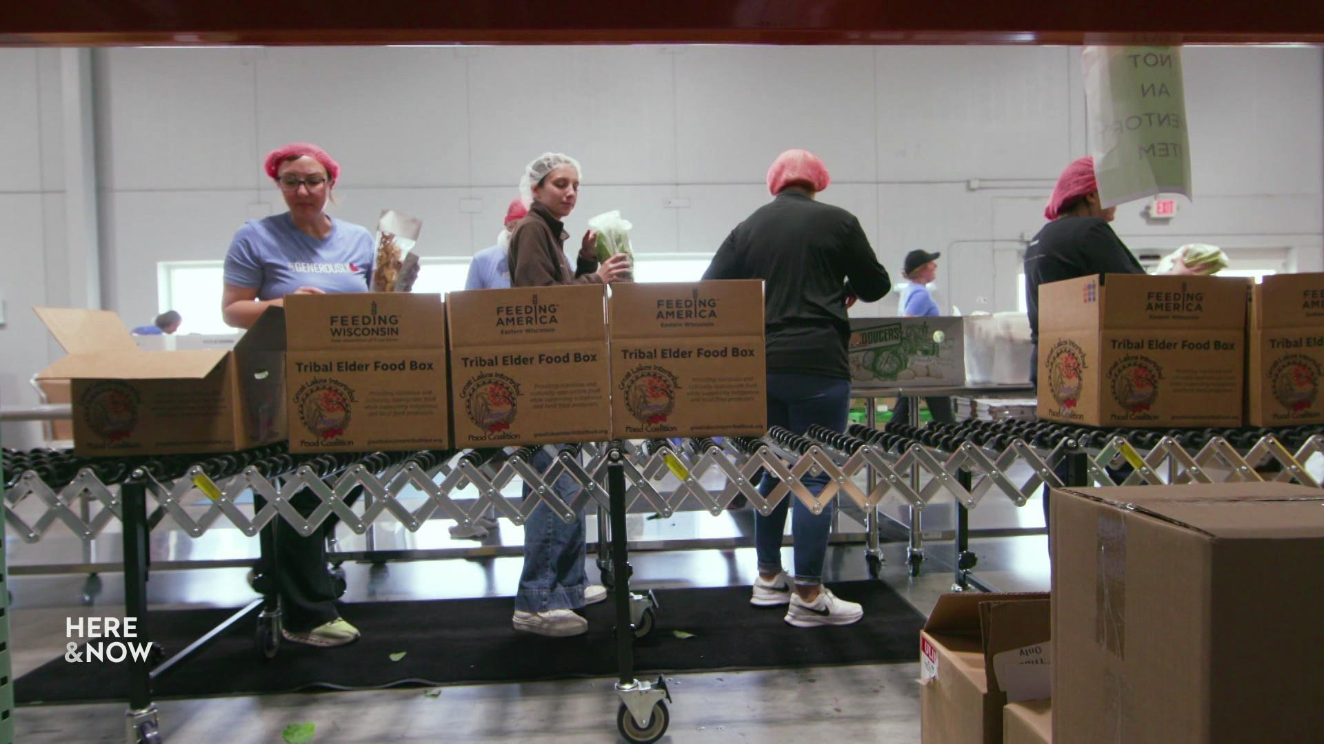 Stacked cardboard boxes labeled Tribal Elder Food Box from the Great Lakes Intertribal Food Coalition, with workers packing boxes in the background.
