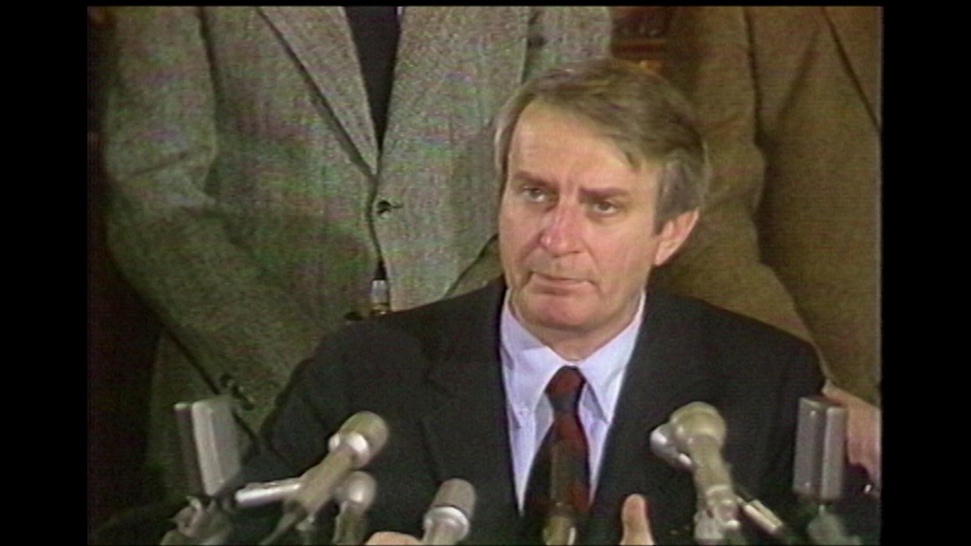 A still image from a video shows Tony Earl siting and speaking into several microphones with other people standing in the background.