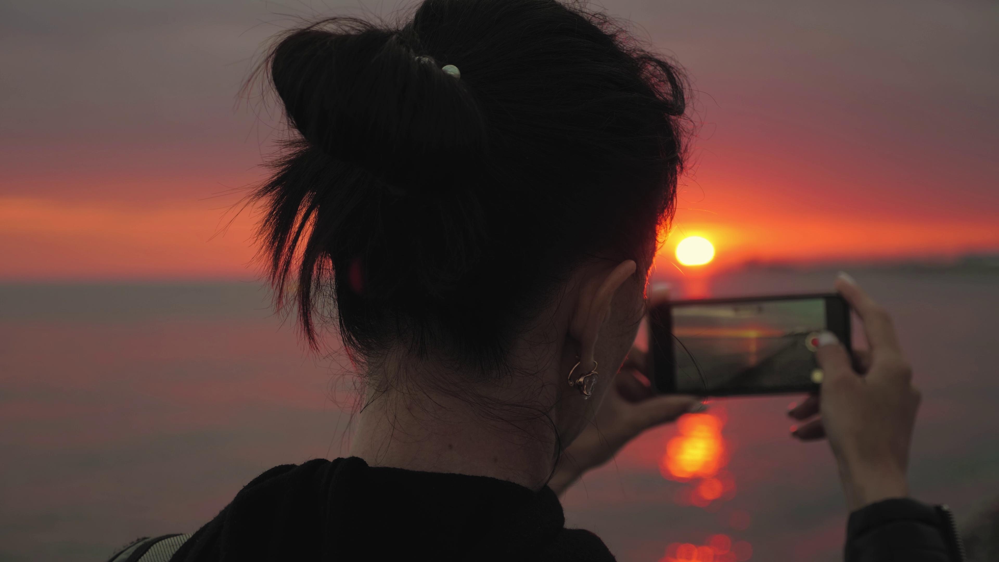 A woman taking an image of the sunset through her phone.
