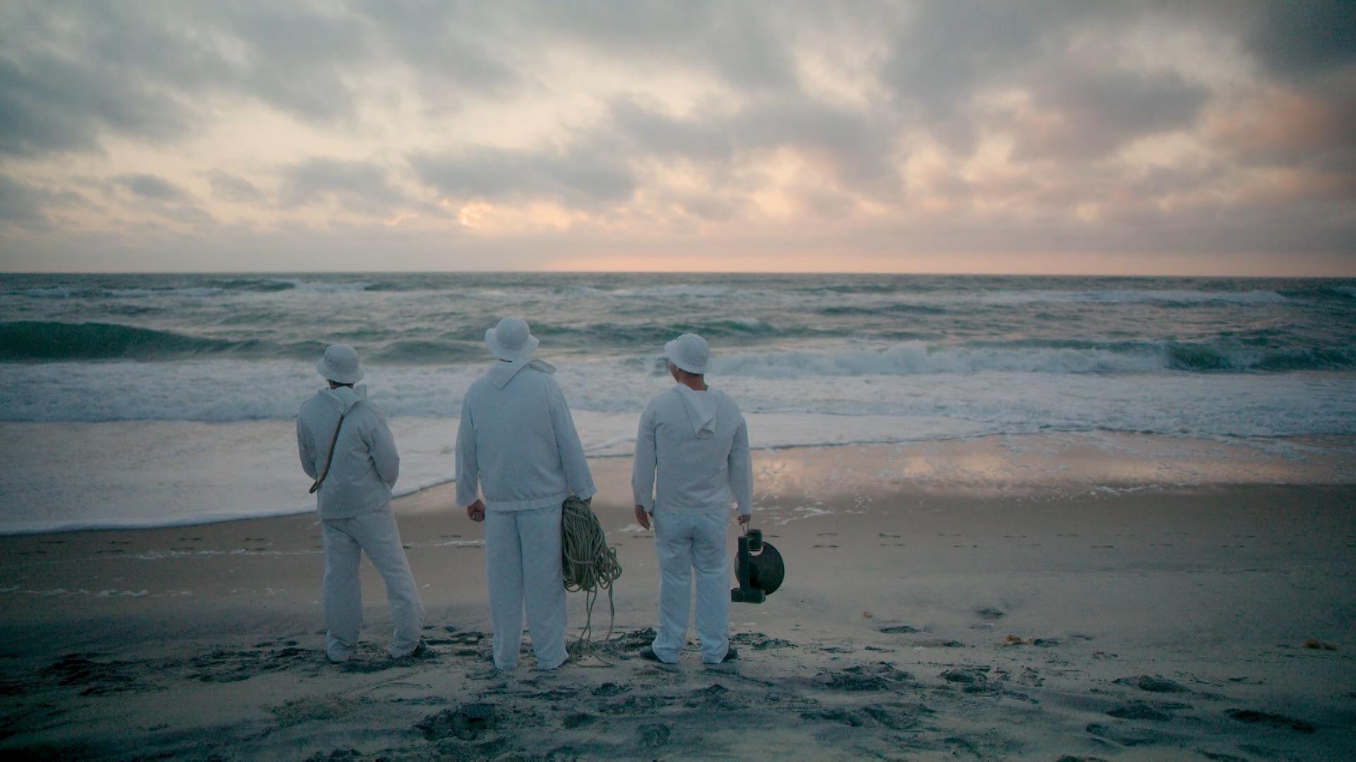 Three people in white suits looking out on the beach horizon.