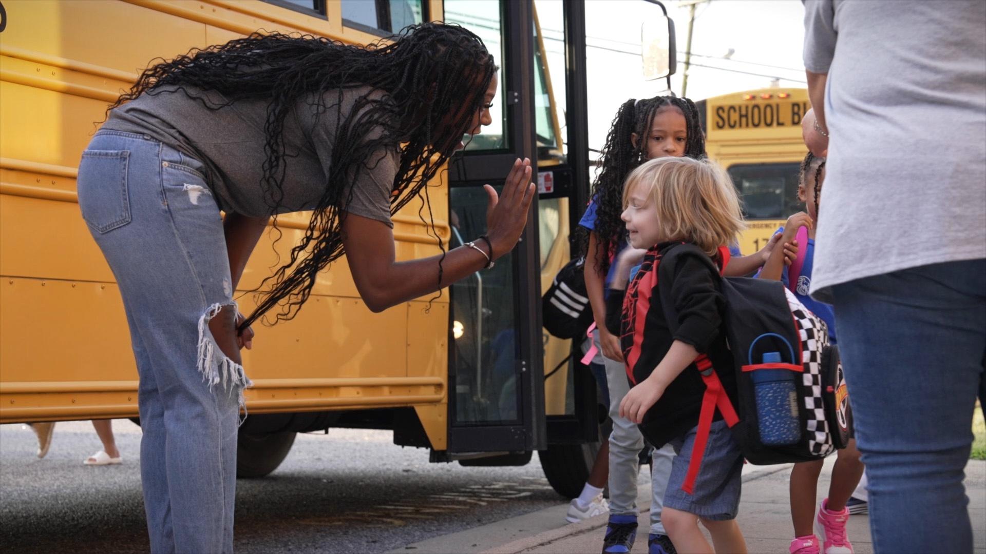A Black woman leaning down to high-five a young child in front of a yellow school bus.