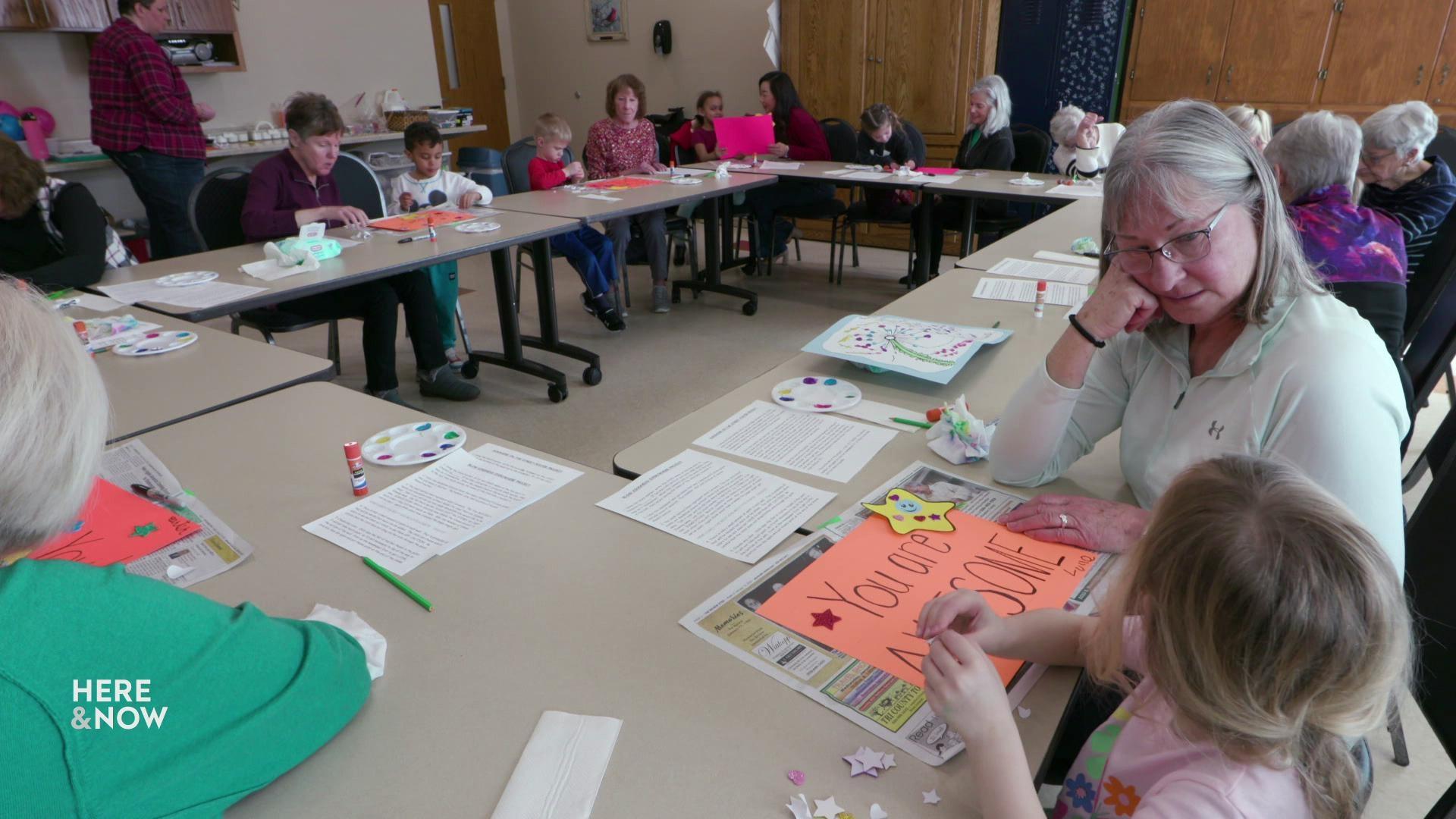 A still image shows a group of older adults and young children doing art and seated in the round at tables
