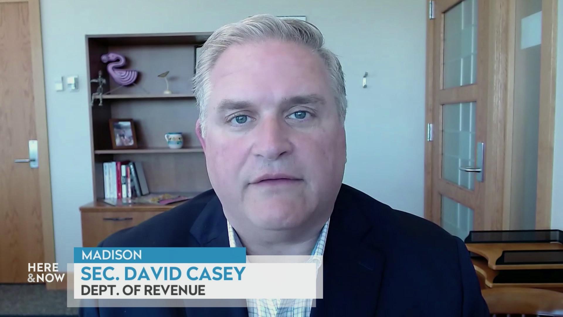 A still image from a video shows David Casey seated in front of a wooden bookshelf with books and items with a graphic at bottom reading 'Madison,' 'David Casey' and 'Dept. of Revenue'