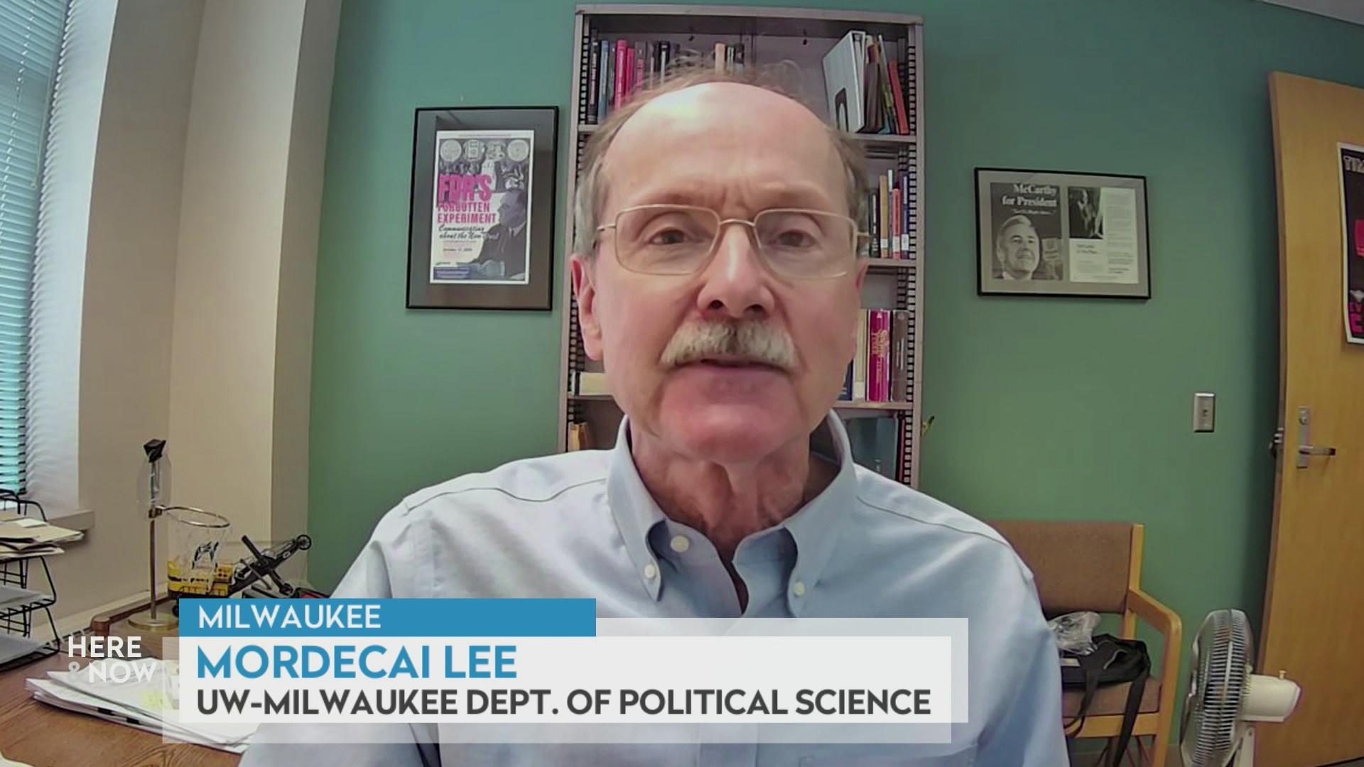 A still image from a video shows Mordecai Lee seated in front of framed art hanging on the wall on either side with a graphic at bottom reading 'Milwaukee,' 'Mordecai Lee' and 'UW-Milwaukee Dept. of Political Science.'