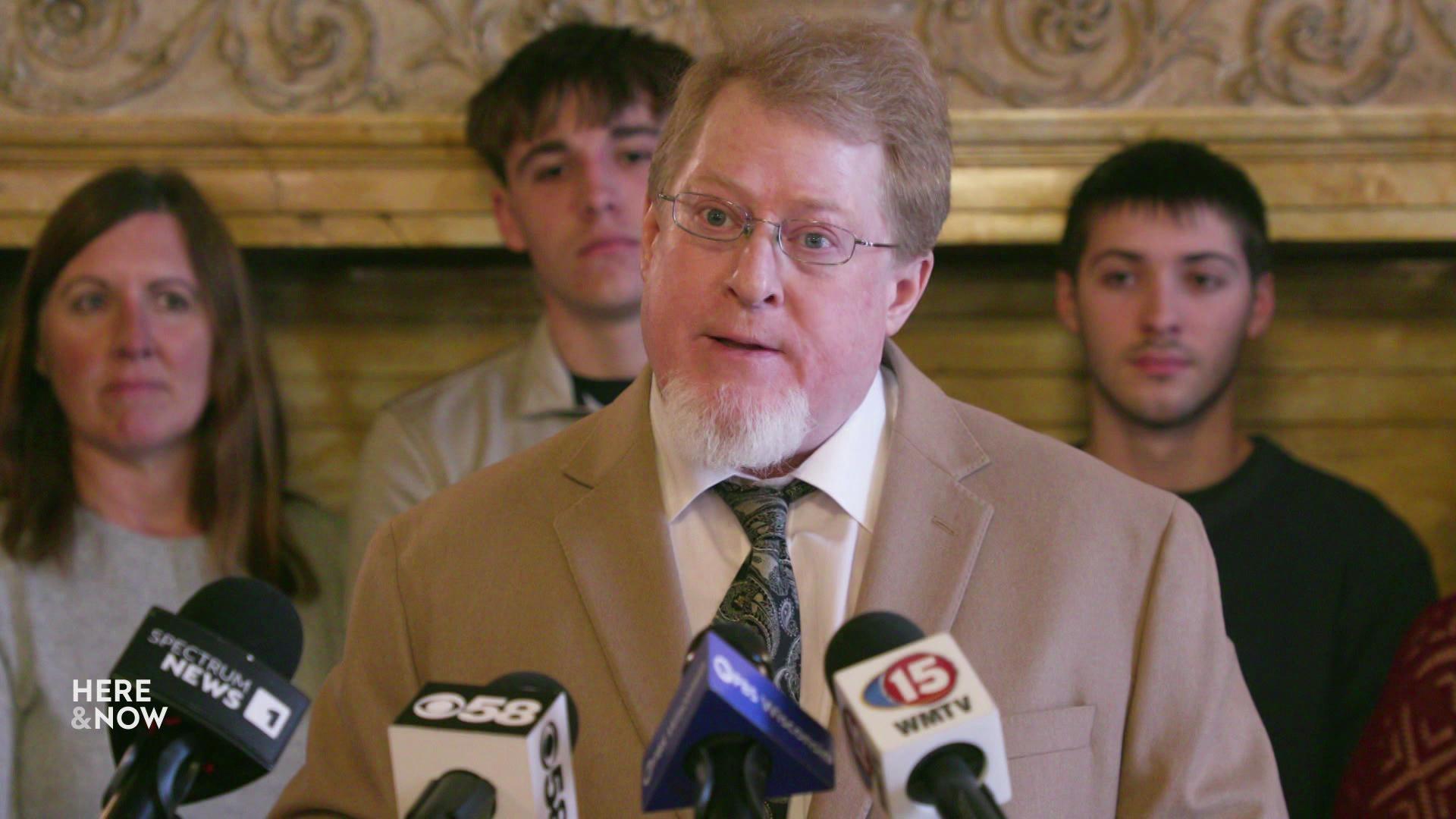 A still image shows Rob Gundermann standing at the state capitol in front of a podium with many mics on it.