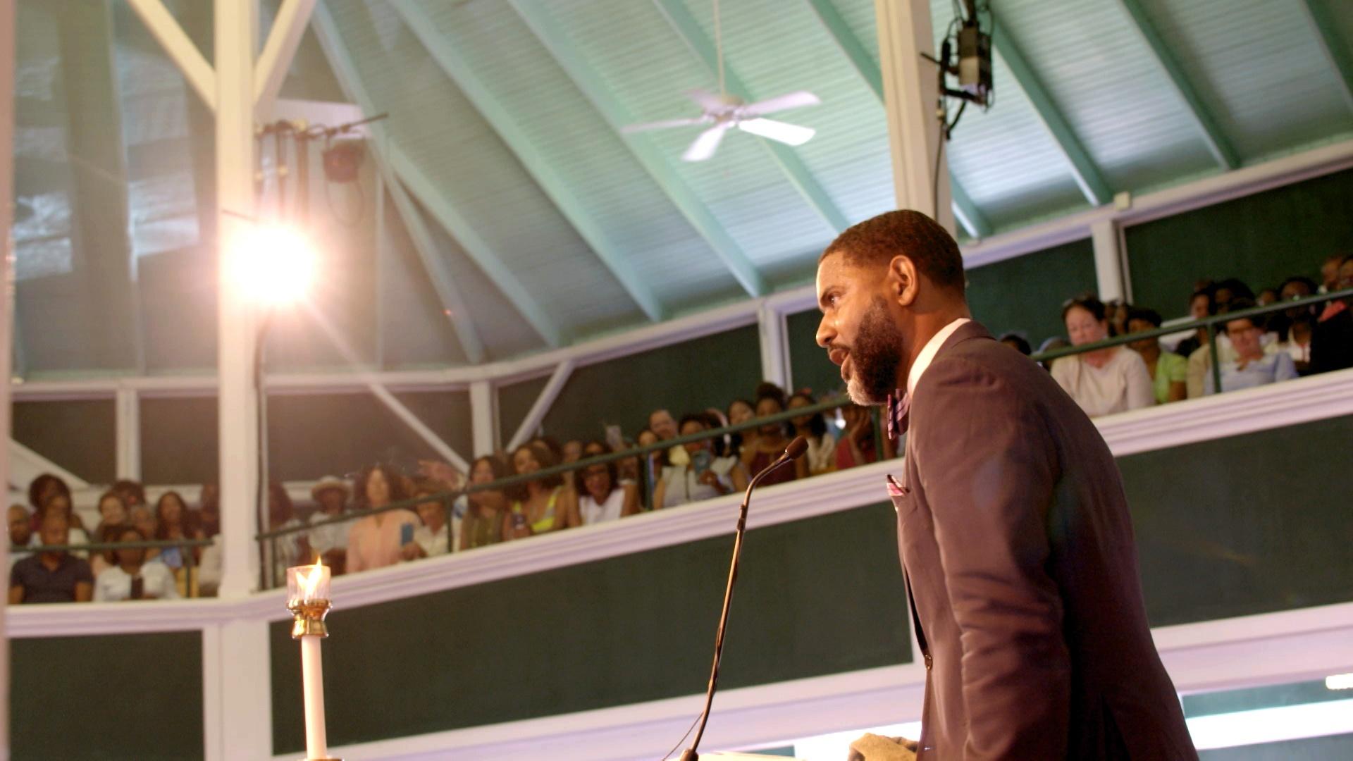 A young man in a suit with a bowtie and pocket square stands at a microphone with a crowded church behind him