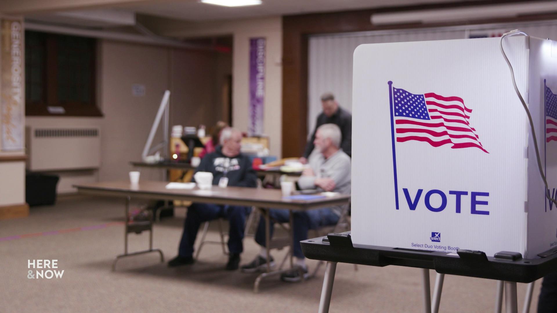 A still image shows two people seated at a table out of focus with a polling booth with a screen with the word 'Vote' and a graphic of the U.S. flag in focus to the right.