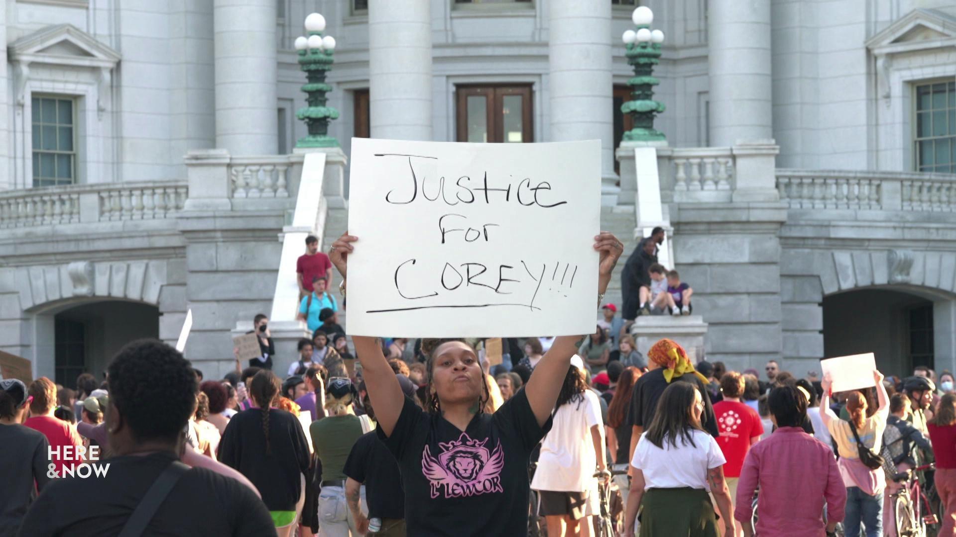A still image shows a crowd of people marching on the steps of the state Capitol, with a woman wearing a black graphic t-shirt in the foreground holding a sign that reads 'Justice for COREY!!!