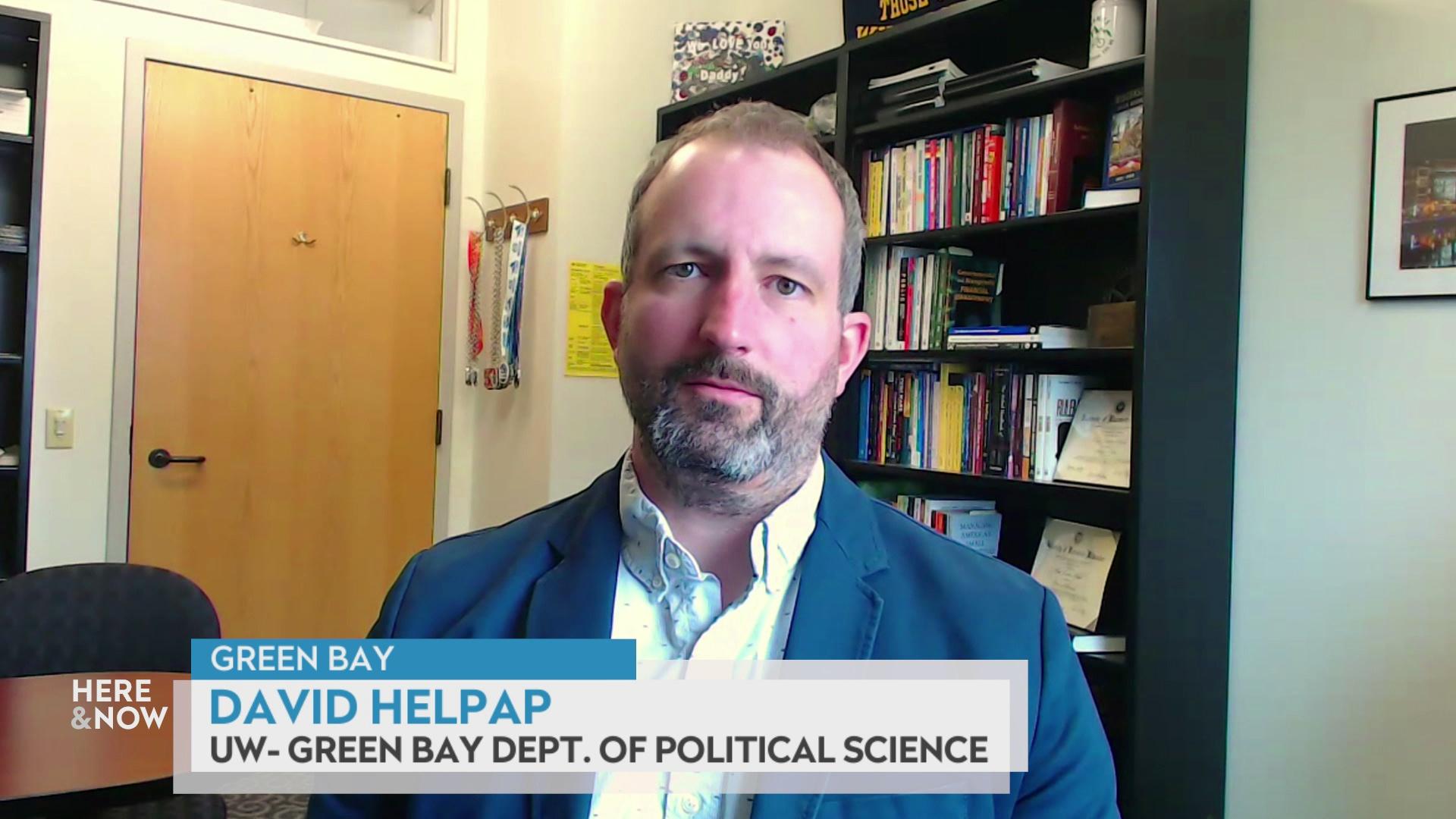 A still image from a video shows David Helpap seated in front of a bookshelf and wooden door with a graphic at bottom reading 'Green Bay,' 'David Helpap' and 'UW-Green Bay Dept. of Political Science.'