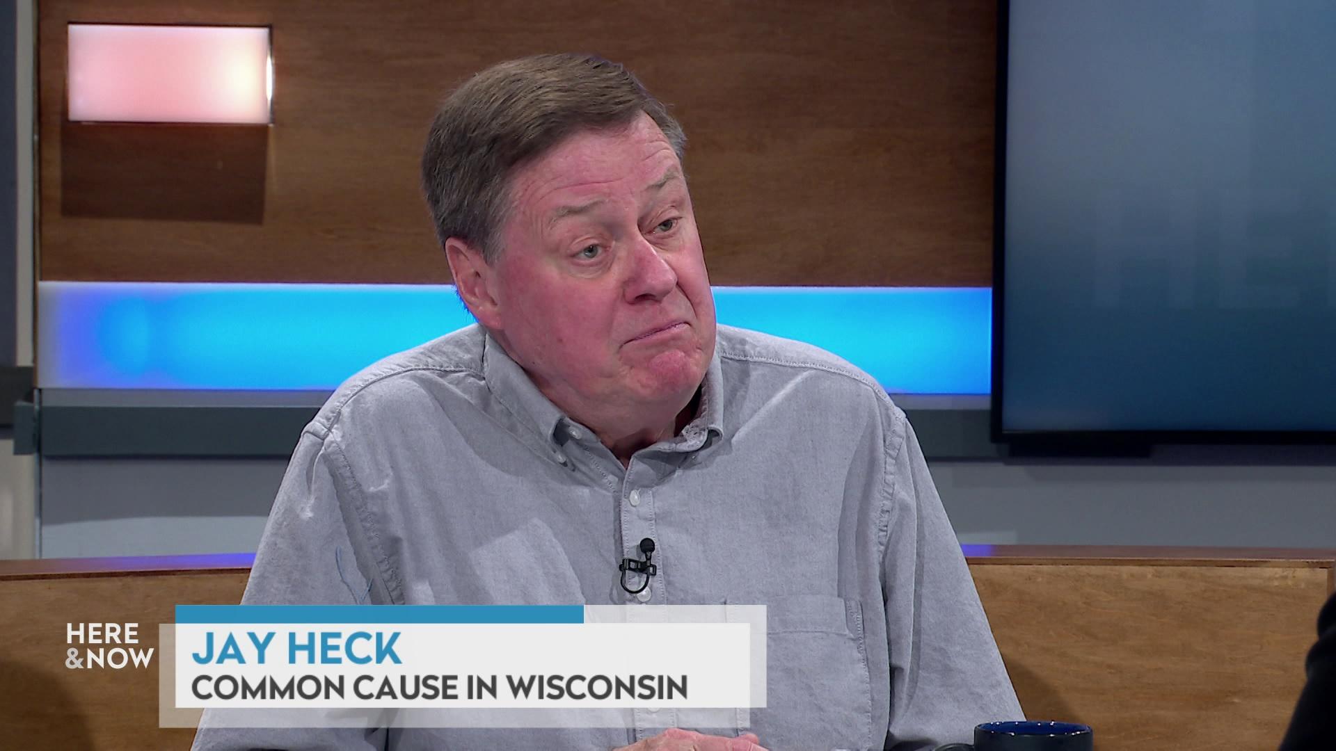 A still image shows Jay Heck seated at the 'Here & Now' set featuring wood paneling, with a graphic at bottom reading 'Jay Heck' and 'Common Cause in Wisconsin.'
