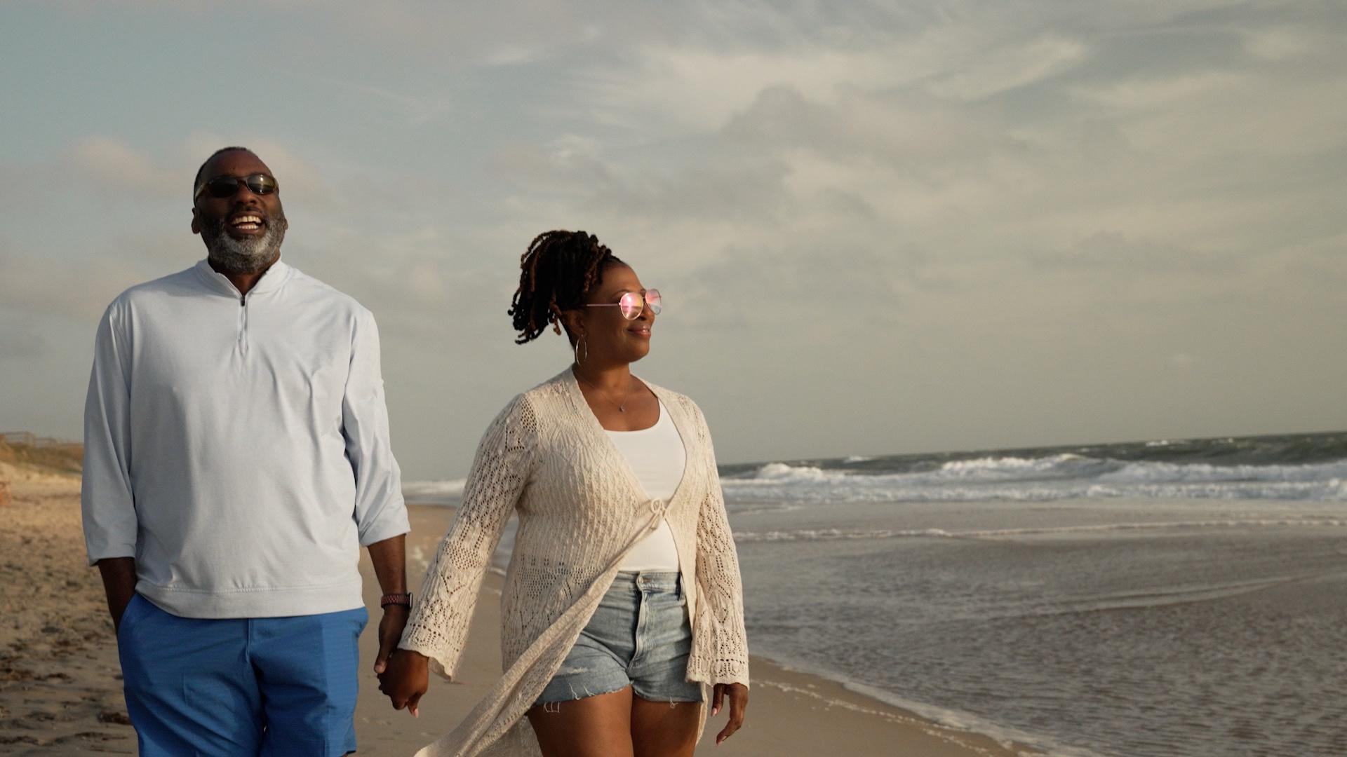 A Black man and a Black woman holding hands and smiling while walking on the beach.
