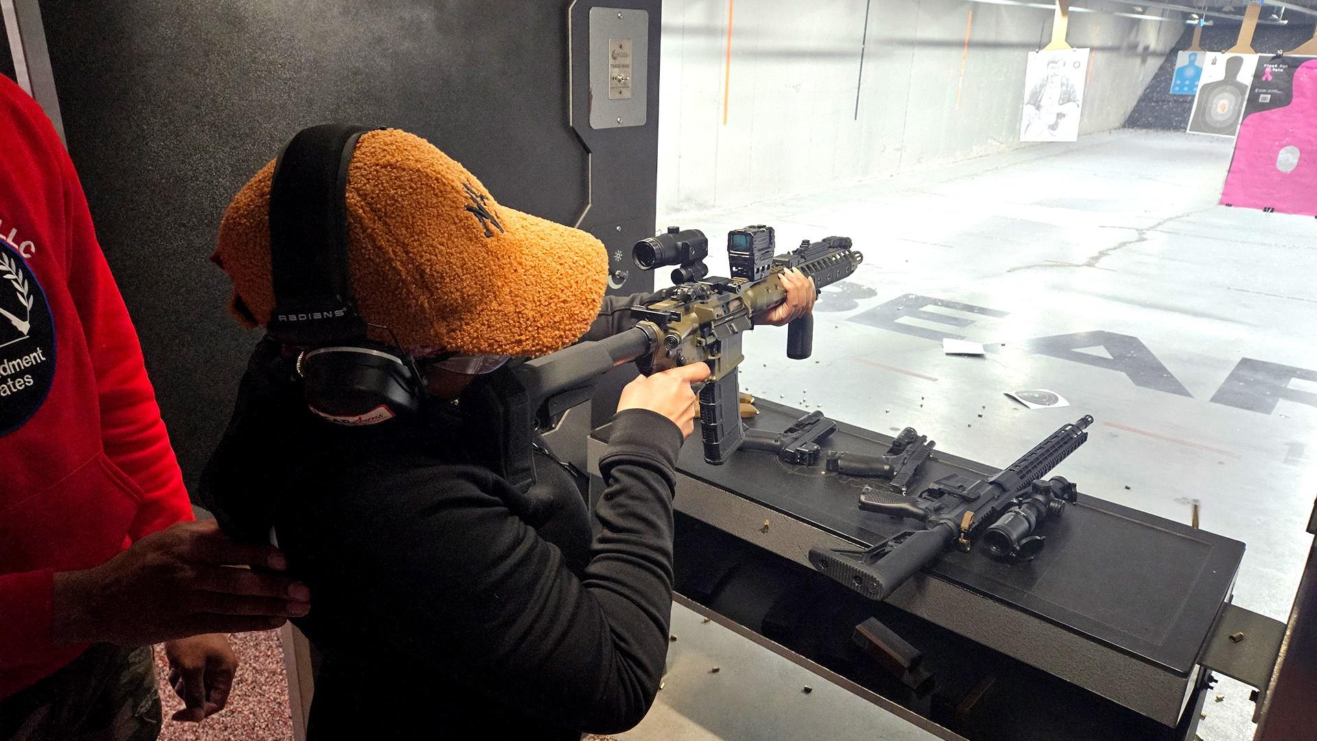 Person aiming a scoped rifle at a target in an indoor shooting range, with firearms and gear on the bench nearby.