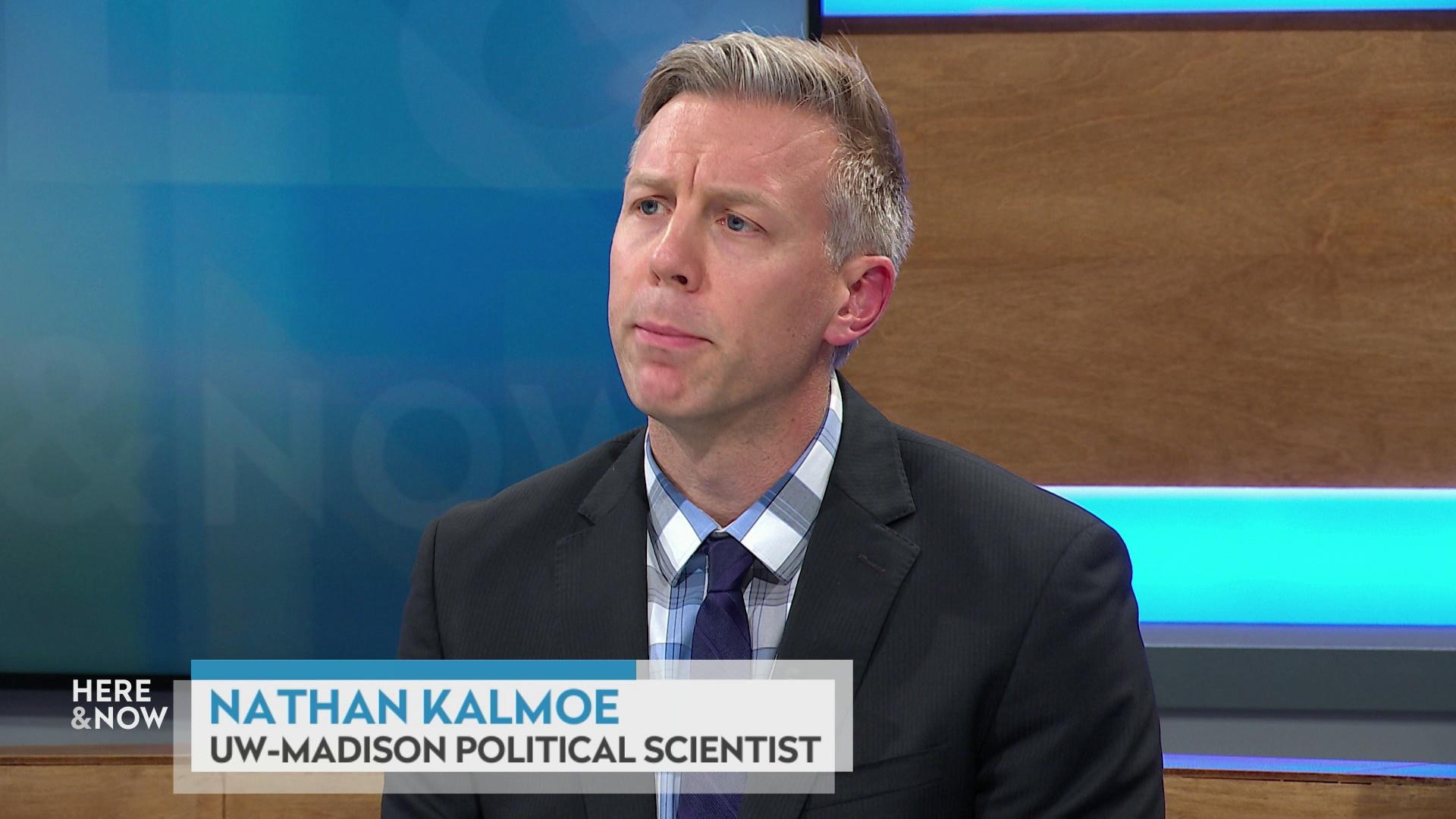 A still image shows Nathan Kalmoe seated at the 'Here & Now' set featuring wood paneling, with a graphic at bottom reading 'Nathan Kalmoe' and 'UW-Madison Political Scientist.'