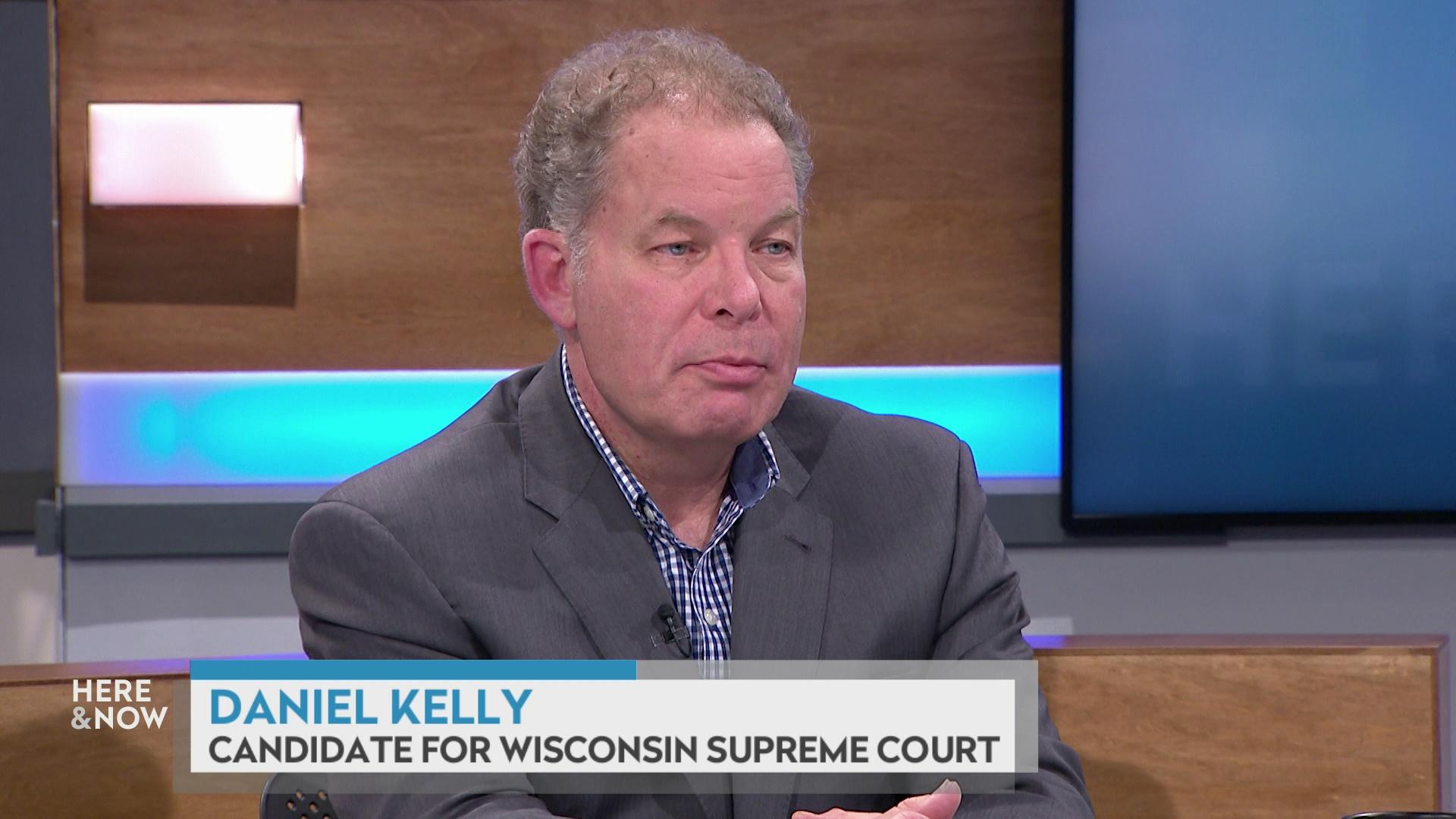 A still image from a video shows Daniel Kelly seated at the 'Here & Now' set featuring wood paneling, with a graphic at bottom reading 'Daniel Kelly' and 'Wisconsin Supreme Court Candidate.'