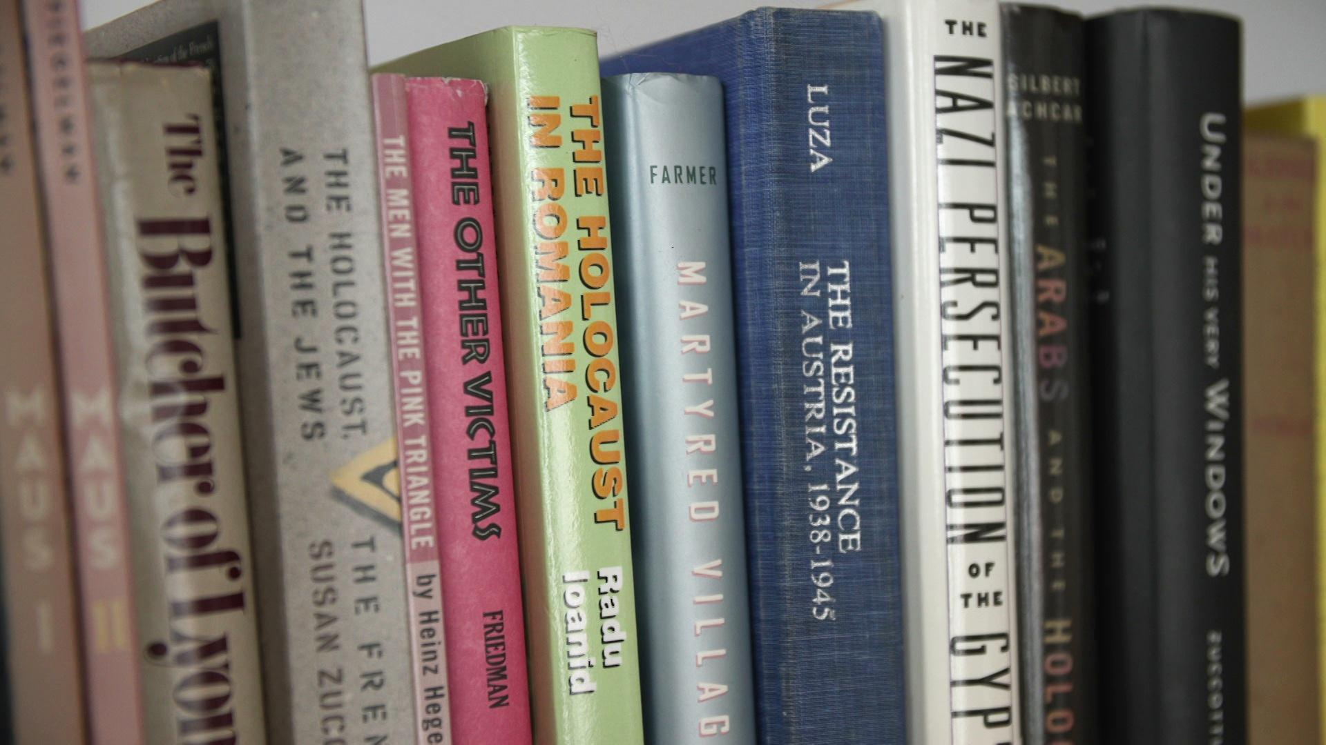 A still image from a video shows a row of books about Jewish history and the Holocaust on a shelf, including titles that read 'The Other Victims,' 'The Holocaust in Romania' and 'The Resistance in Austria, 1938-1945.'