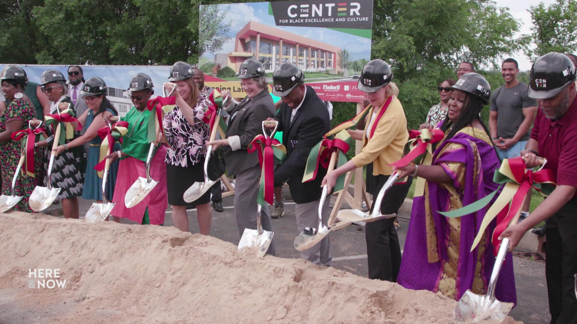 A line of people wearing hardhats and holding shovels scoop up mounds of dirt in front of them.