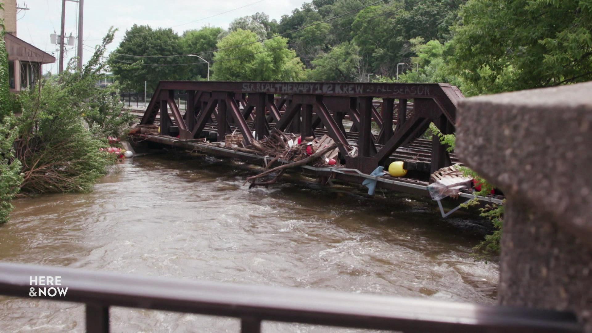 A still image shows a railroad bridge in Milwaukee with logs and debris interwoven in its bars, it spans across a river of unusually high water.