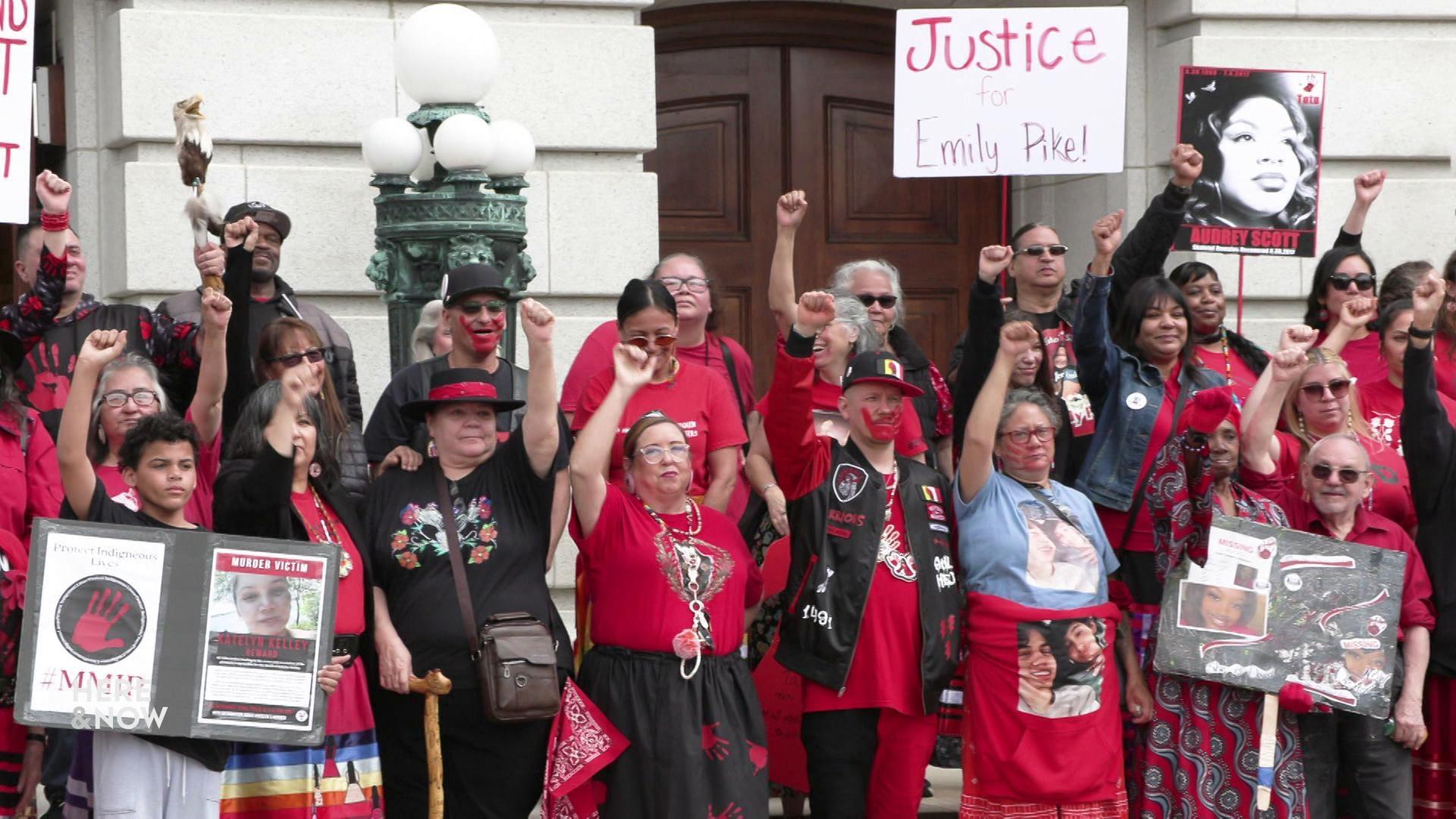 A large group wearing red and black stand in front of the capital holding their fists and signs in the air