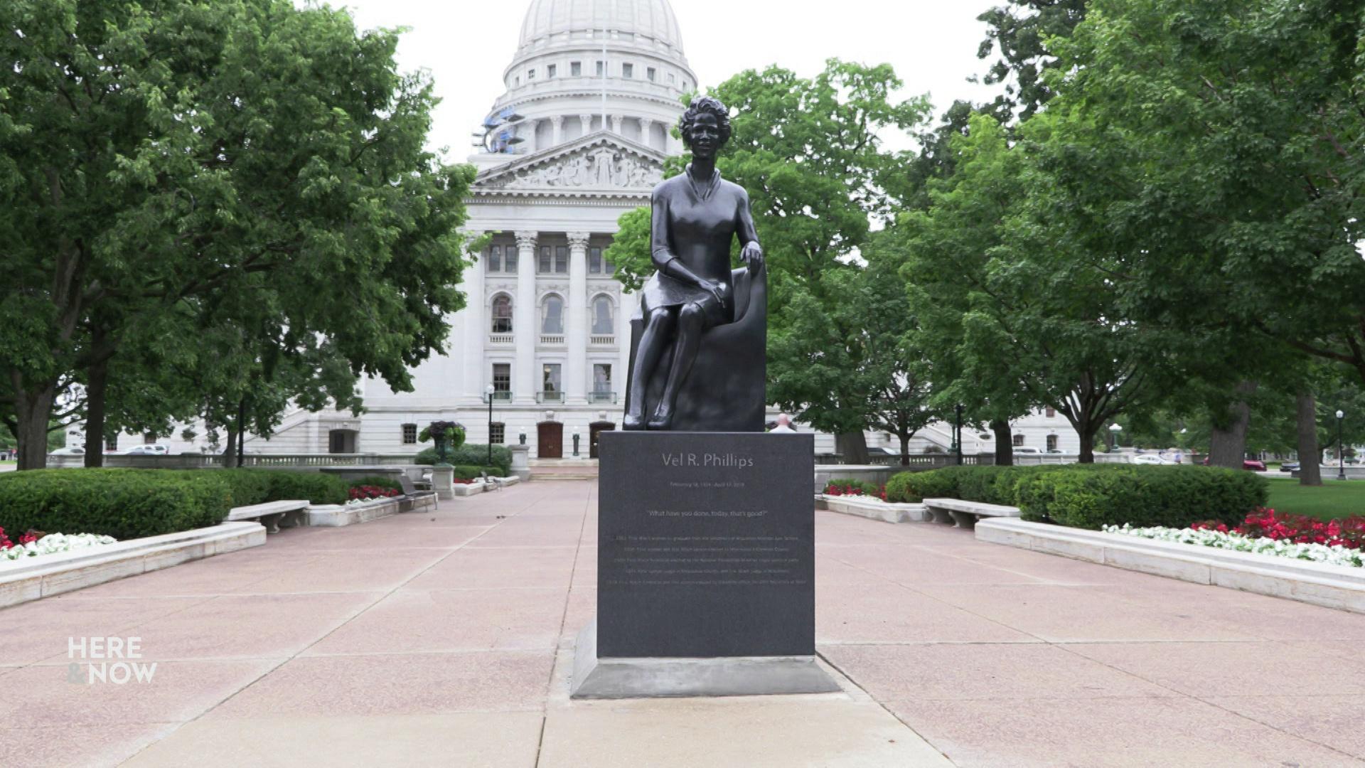 A still image shows a statue depicting Vel Phillips on cement in front of the Wisconsin State Capitol with trees and flowerbeds in the background.