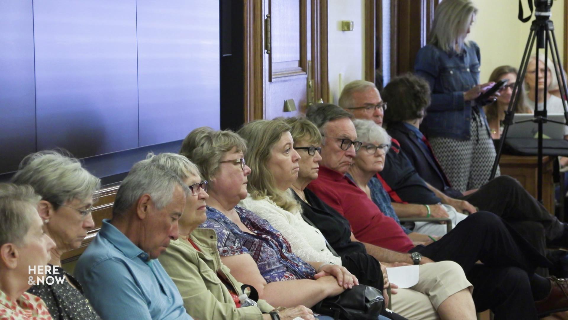 A row of people remain seated along a wall and stare out into a conference room.