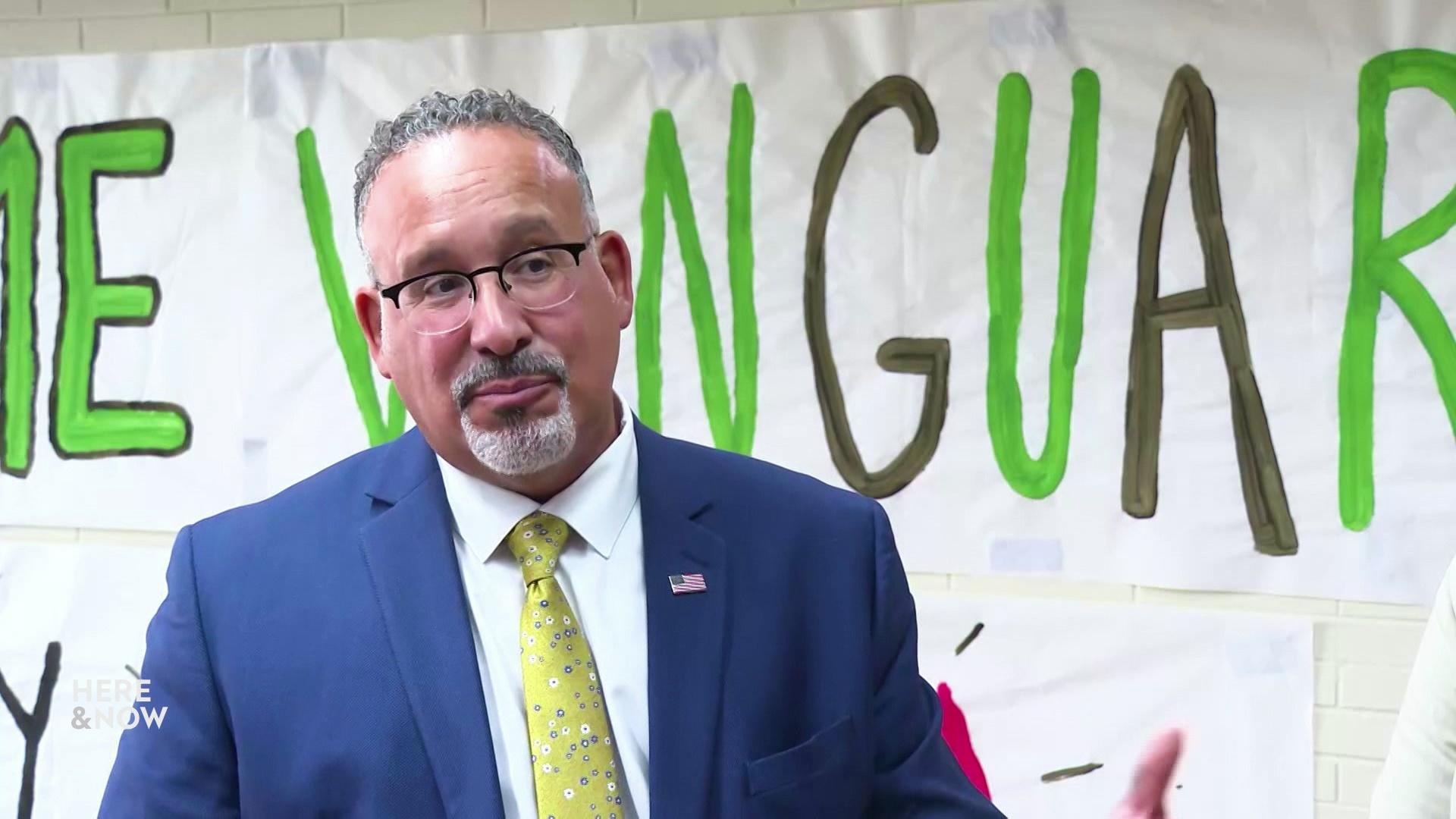 Miguel Cardona speaks and stands in front of a paper poster with painted lettering.