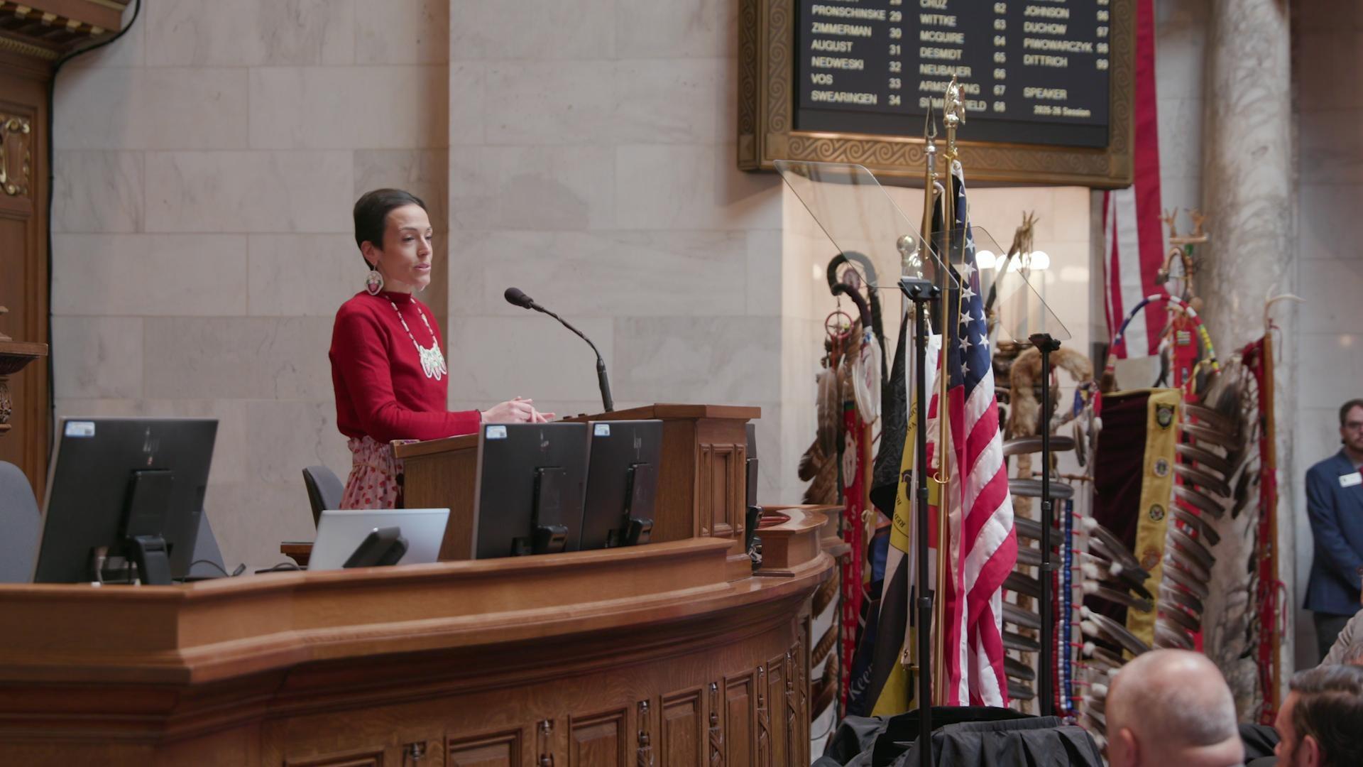 A still image shows a procession of people carrying various Tribal flags walking down the aisle of the State legislature. A still image shows a procession of people carrying various Tribal flags walking down the aisle of the State legislature.