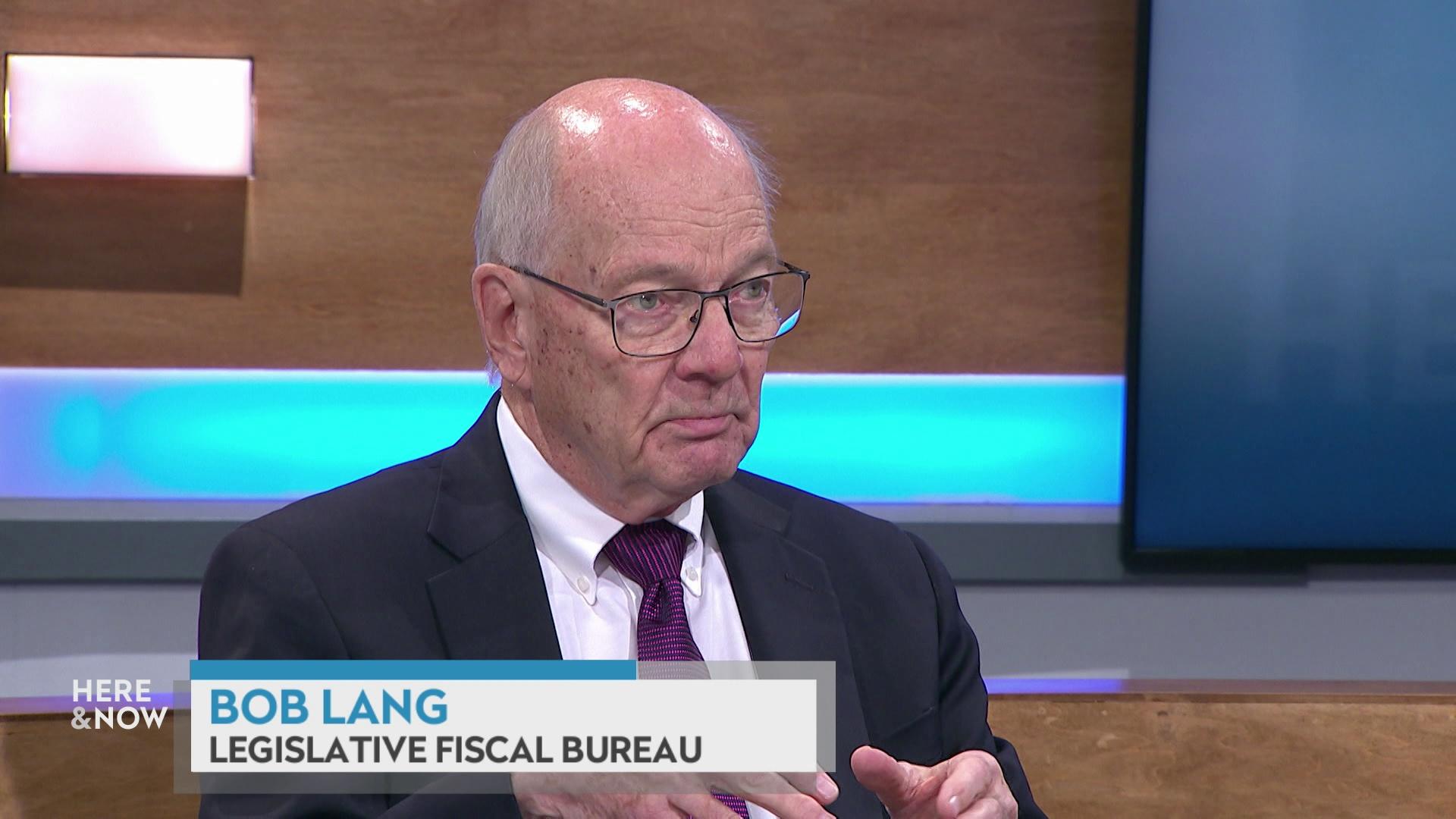 A still image shows Bob Lang seated at the 'Here & Now' set featuring wood paneling, with a graphic at bottom reading 'Bob Lang' and 'Legislative Fiscal Bureau.'