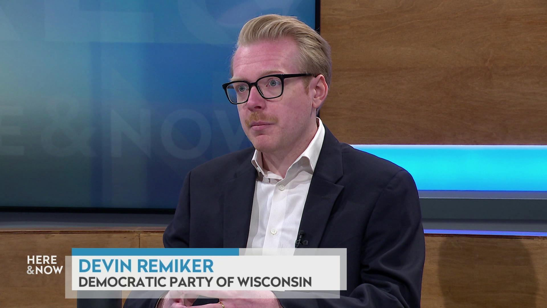 A still image shows Devin Remiker seated at the 'Here & Now' set featuring wood paneling, with a graphic at bottom reading 'Devin Remiker' and 'Democratic Pary of Wisconsin.'