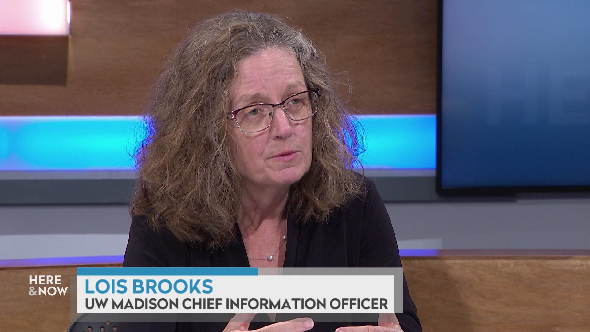 A still image shows Lois Brooks seated at the 'Here & Now' set featuring wood paneling, with a graphic at bottom reading 'Lois Brooks' and 'UW Madison Chief Information Officer.'